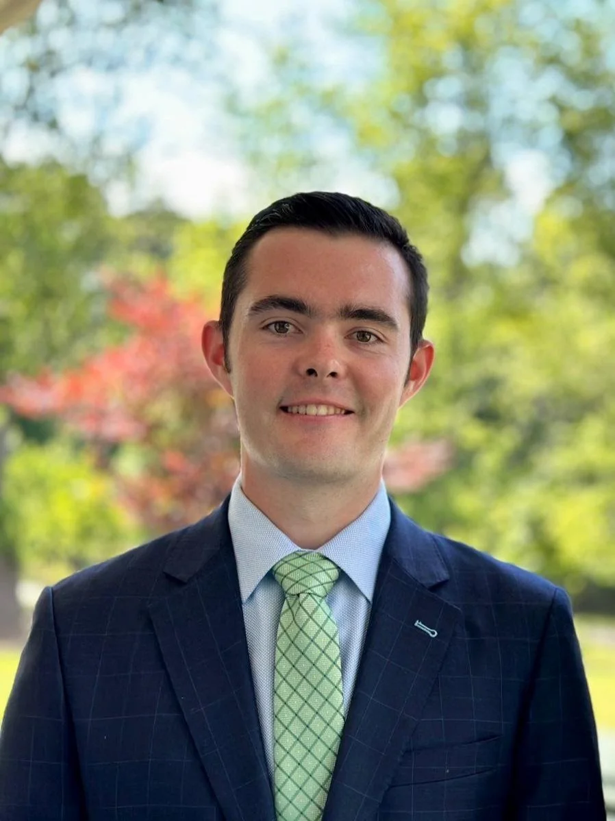 Portrait of a young man in a suit and tie smiling outdoors with trees in the background.