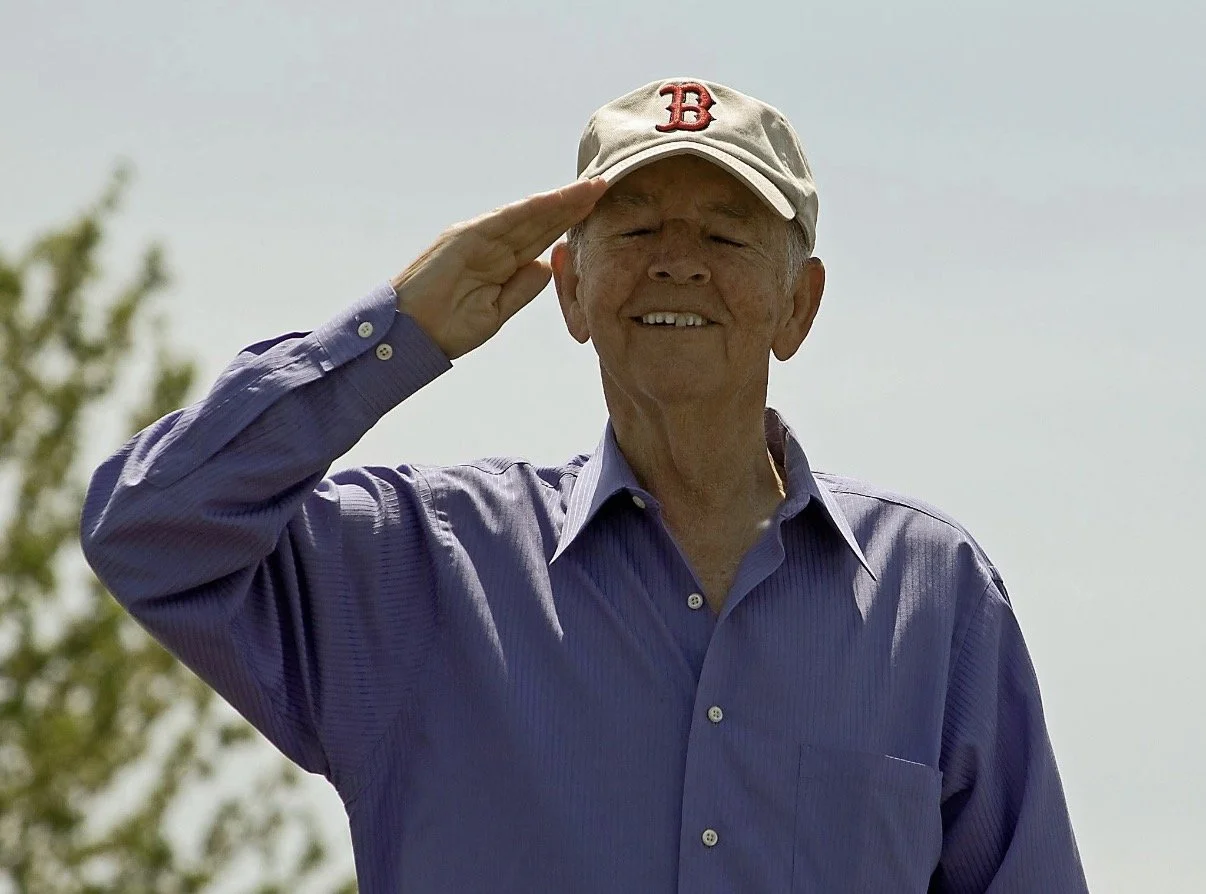 Smiling person in a baseball cap raising a hand in a salute.
