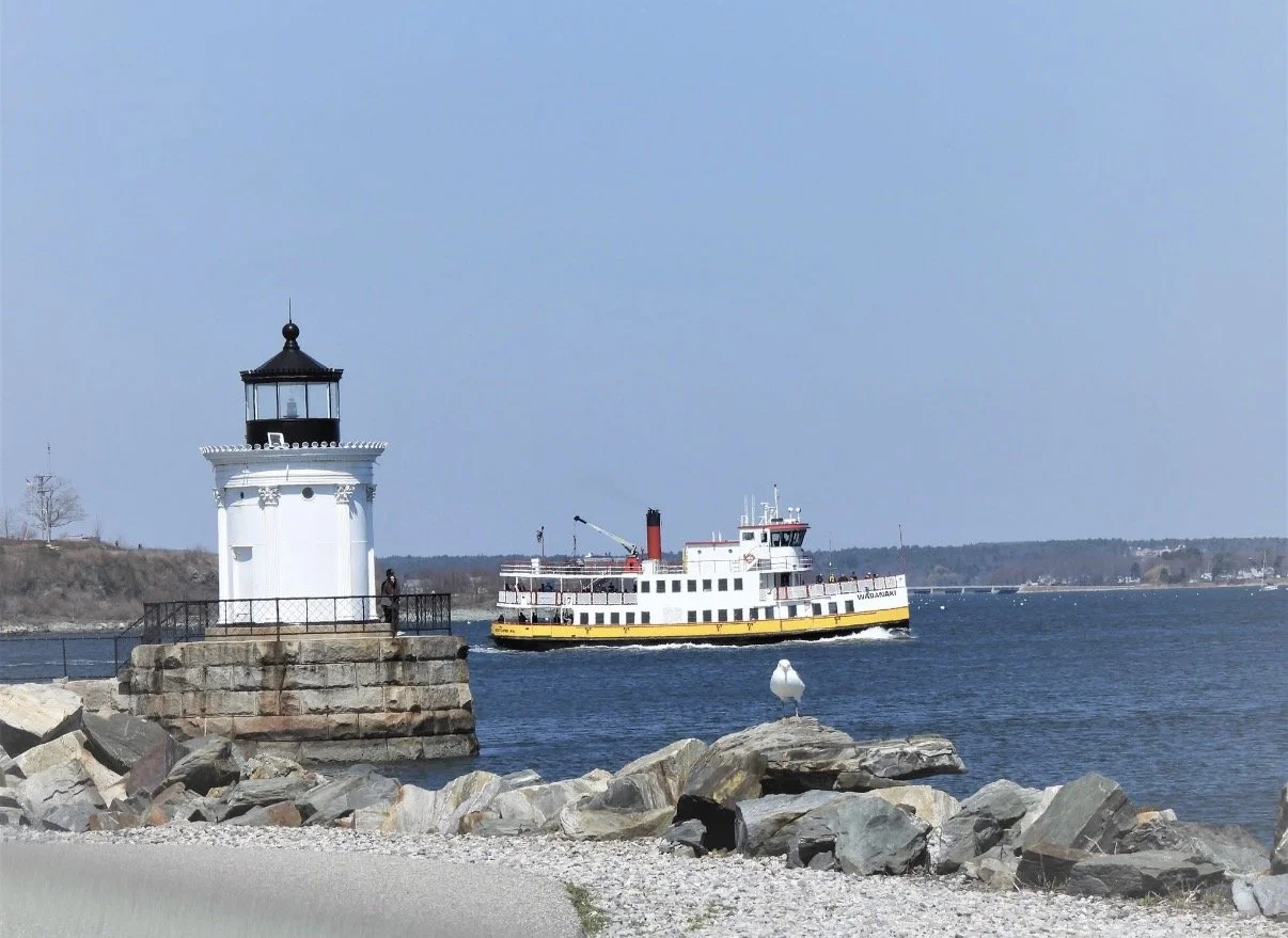 White lighthouse beside a ferry on blue harbor water.