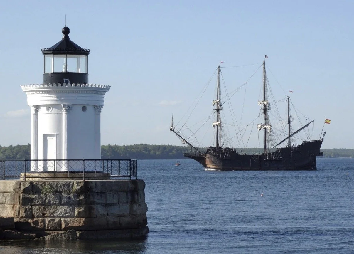 White lighthouse with a tall sailing ship behind it.