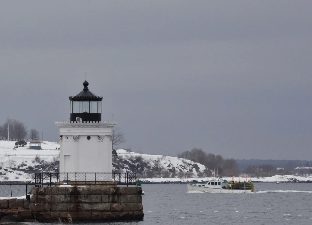 White lighthouse beside a snowy shoreline under gray skies.
