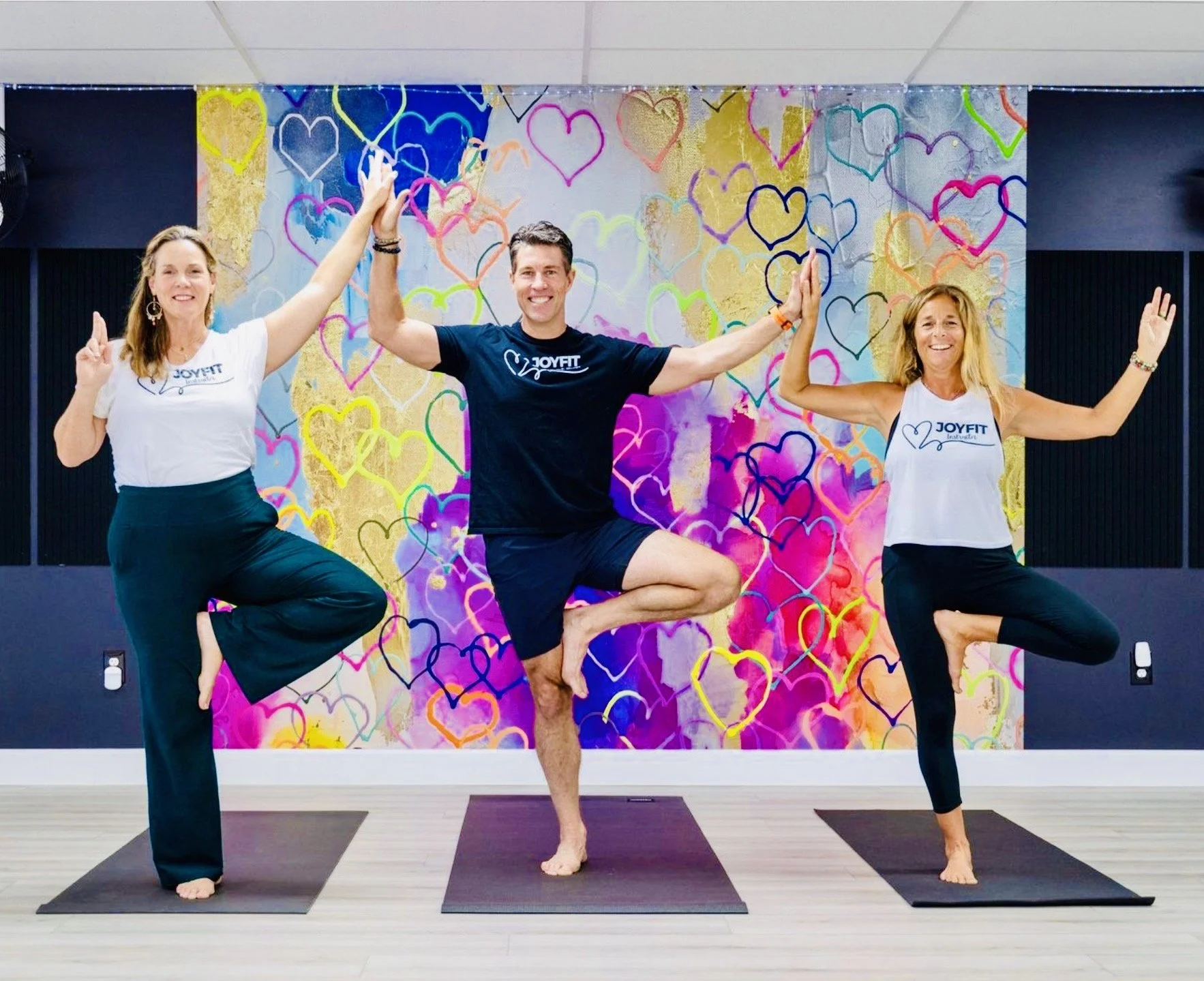 Three people practicing yoga on mats in a studio with a colorful heart-patterned mural background, all in tree pose with one leg bent and foot resting on the inner thigh or calf, hands in prayer or raised.
