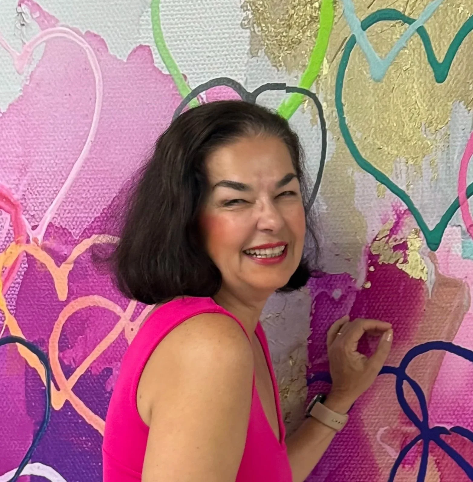 A woman with black hair wearing a sleeveless pink top, smiling and winking, standing in front of a colorful, heart-themed graffiti wall.