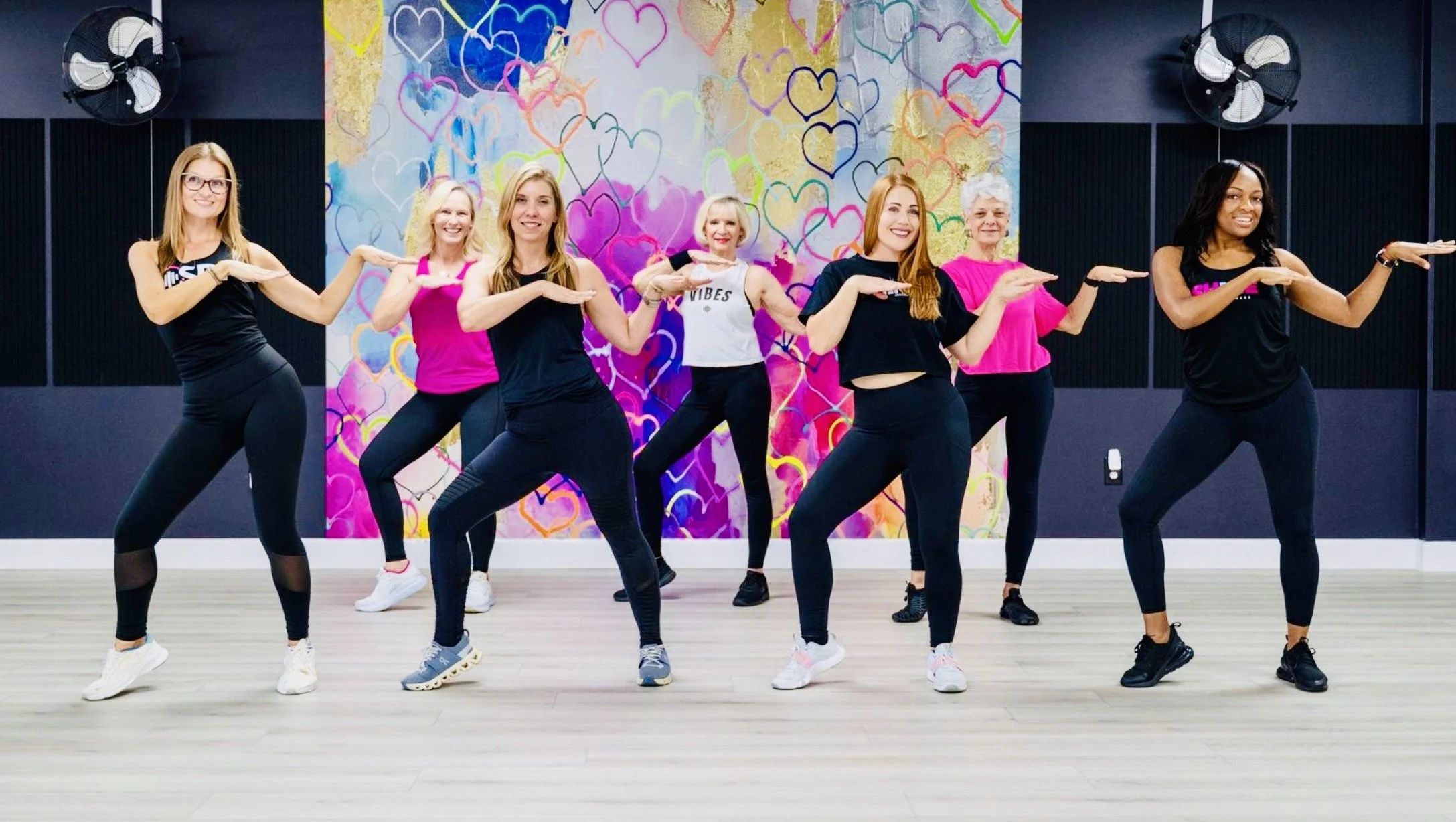 Six women participating in a dance fitness class in a studio with a colorful heart-themed mural background.