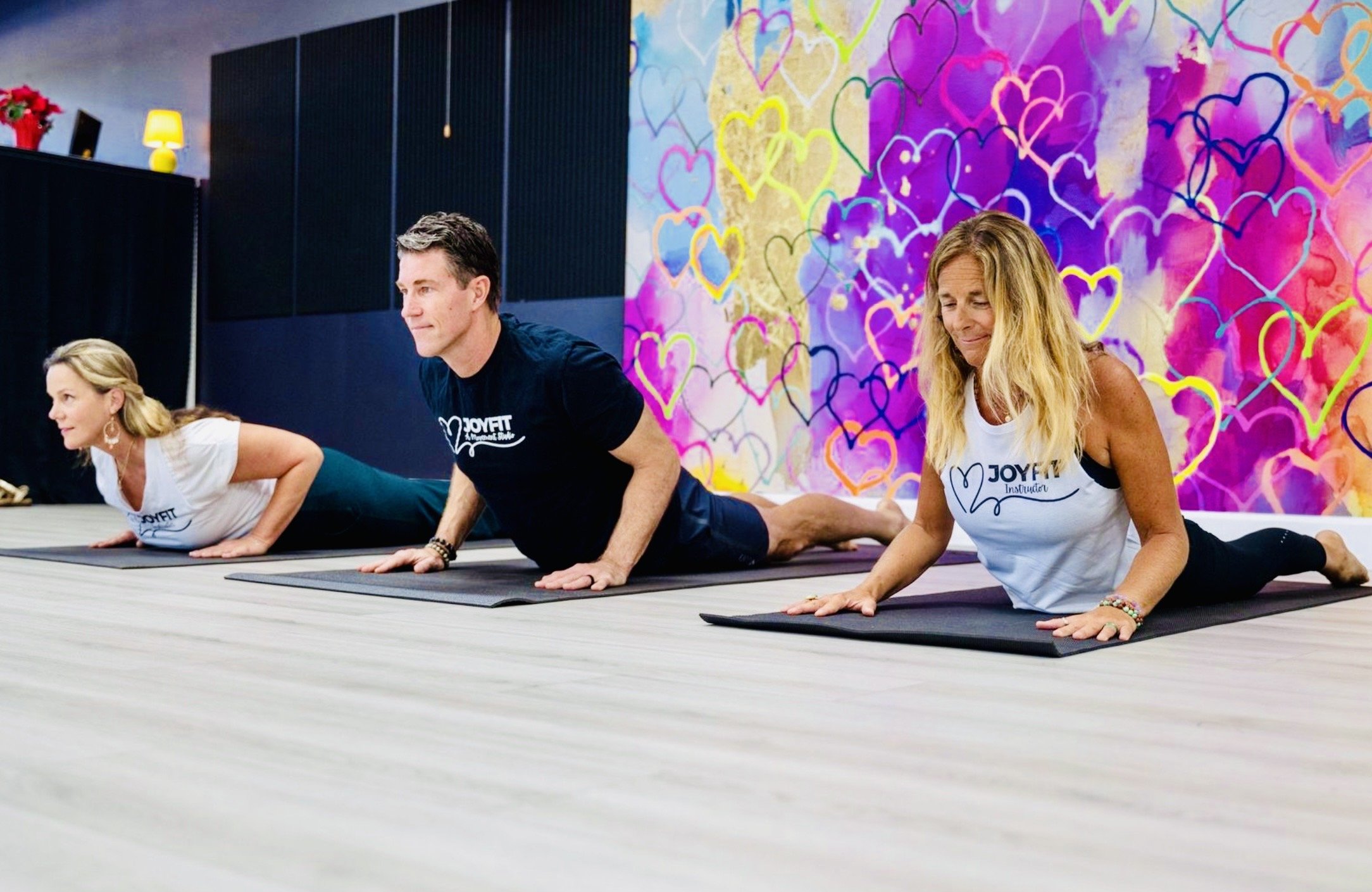 Three people doing yoga in a studio with a colorful heart-themed mural on the wall. They are in a sphinx pose on yoga mats, practicing a gentle backbend.