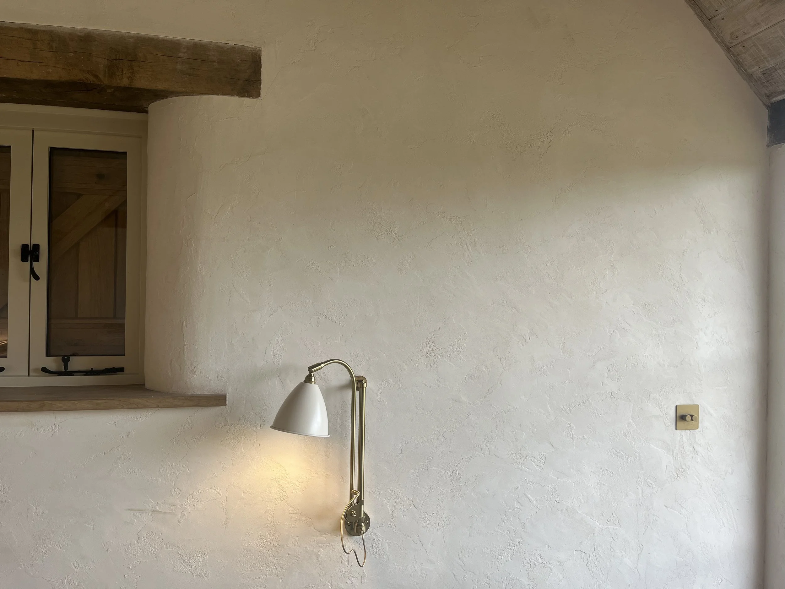 Rustic interior with textured white wall, wooden-framed window, and a brass wall-mounted lamp.