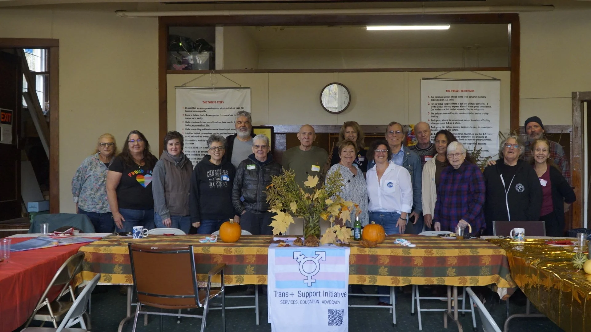 Group of people standing behind a table with pumpkins and autumn leaves, celebrating Trans+ Support Initiative in a community center decorated for fall.