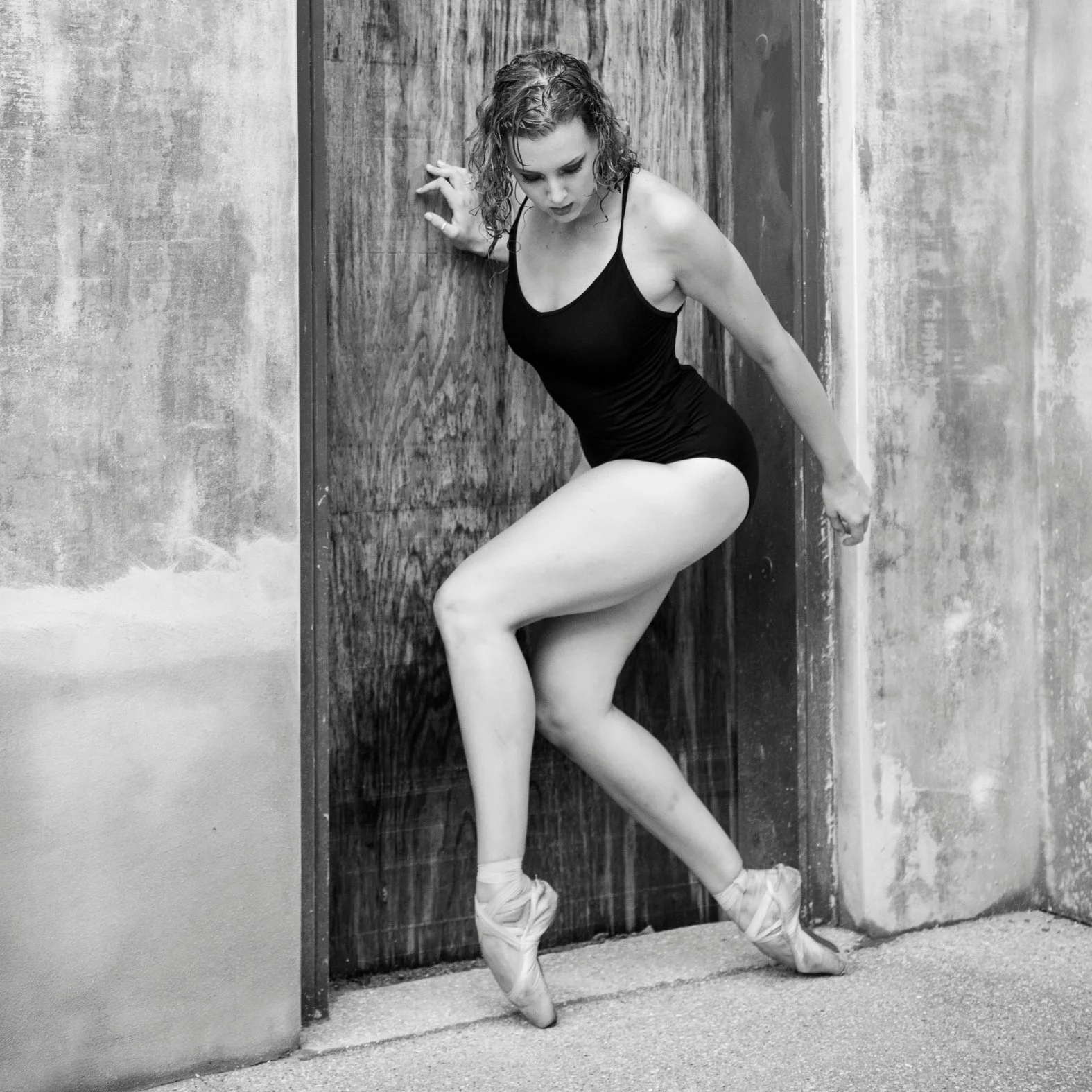 Ballerina posing against a wooden door in a black leotard and pointe shoes