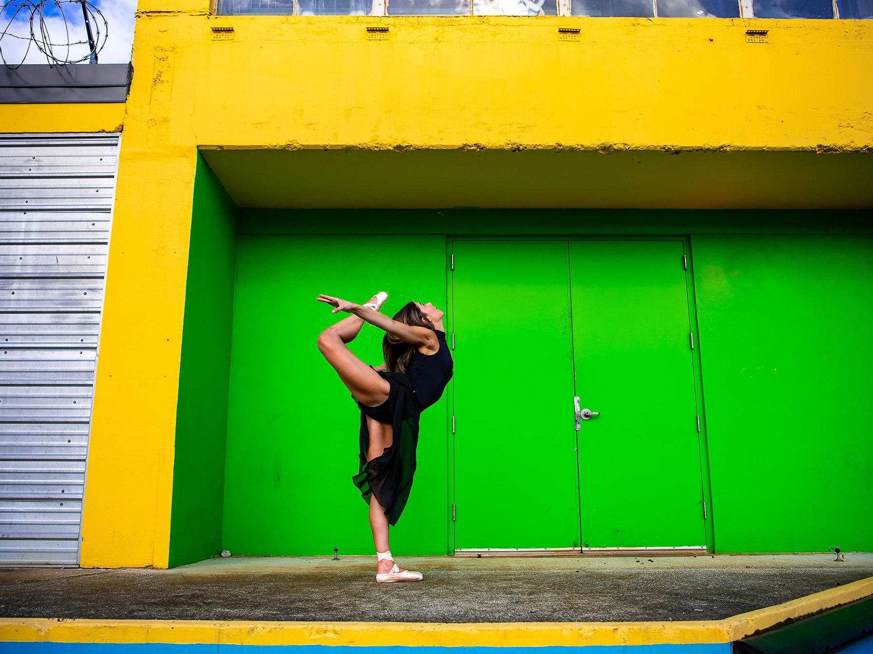 A dancer performs a high leg stretch against a vibrant green and yellow wall background.