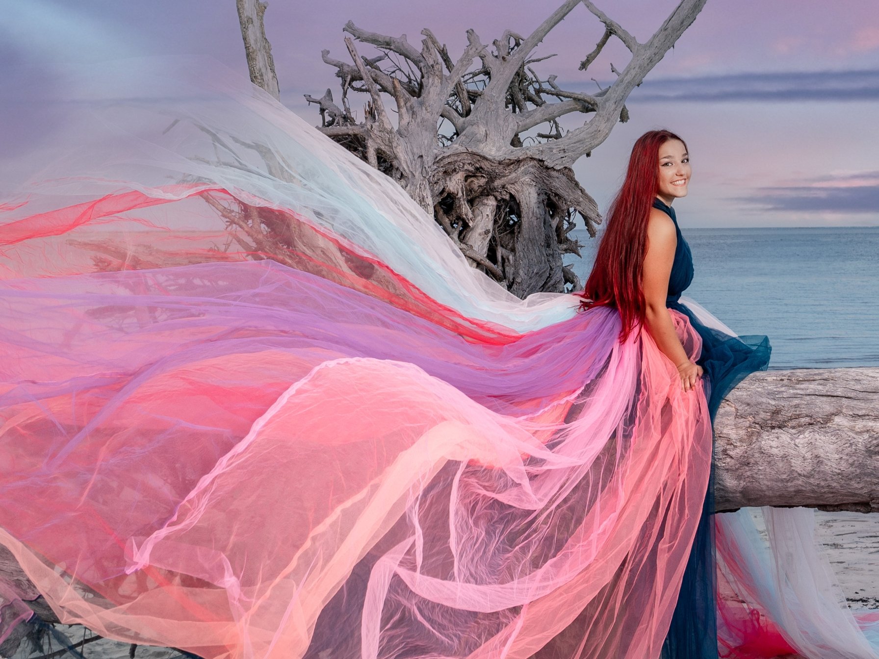 A woman with long red hair sits on a large piece of driftwood by the sea. She wears a dress with a flowing, colorful, translucent train that spreads along the ground. The sky in the background has a soft lavender and blue sunset.