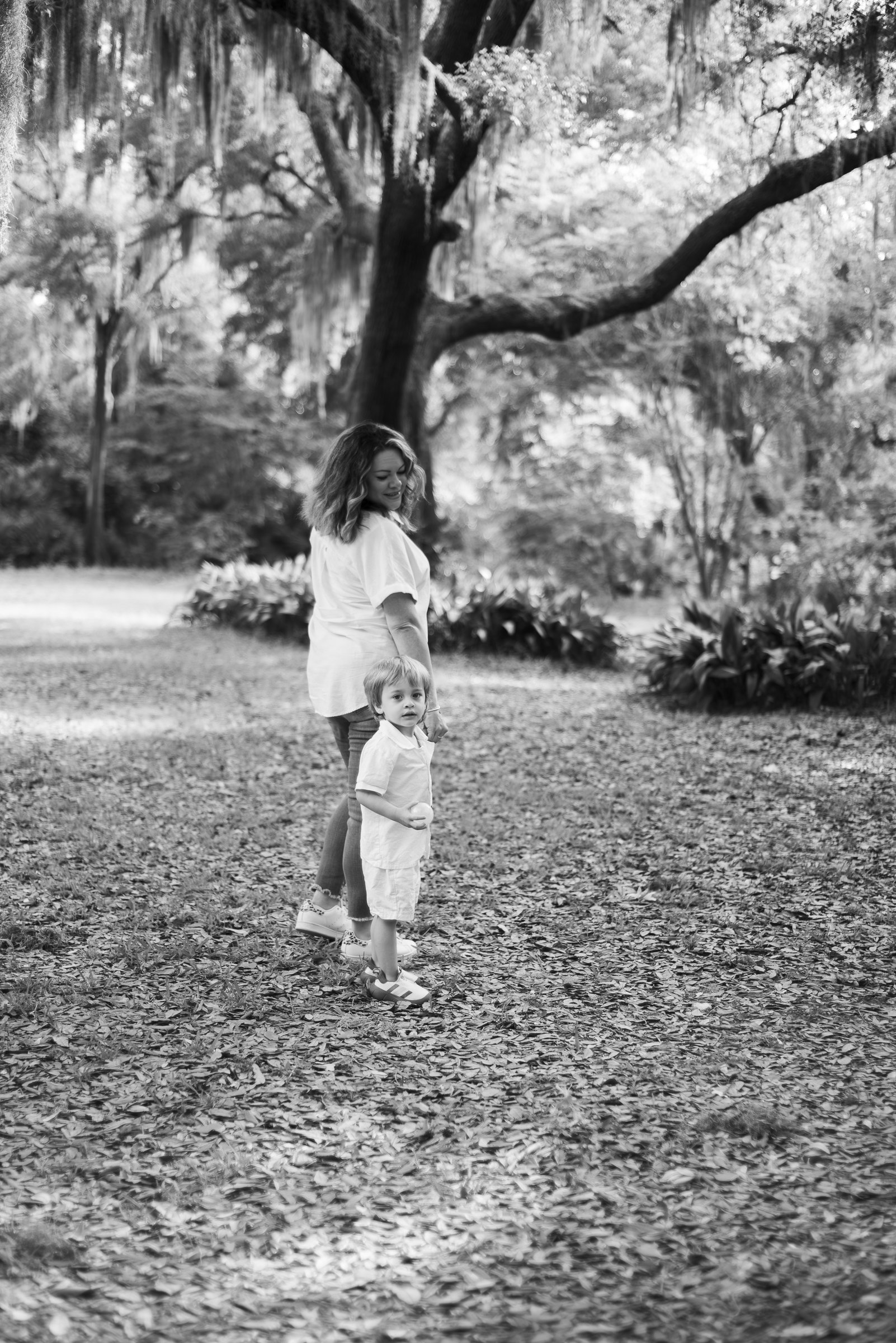 A woman and a child walking in a park, holding hands, surrounded by trees. Black and white photo.
