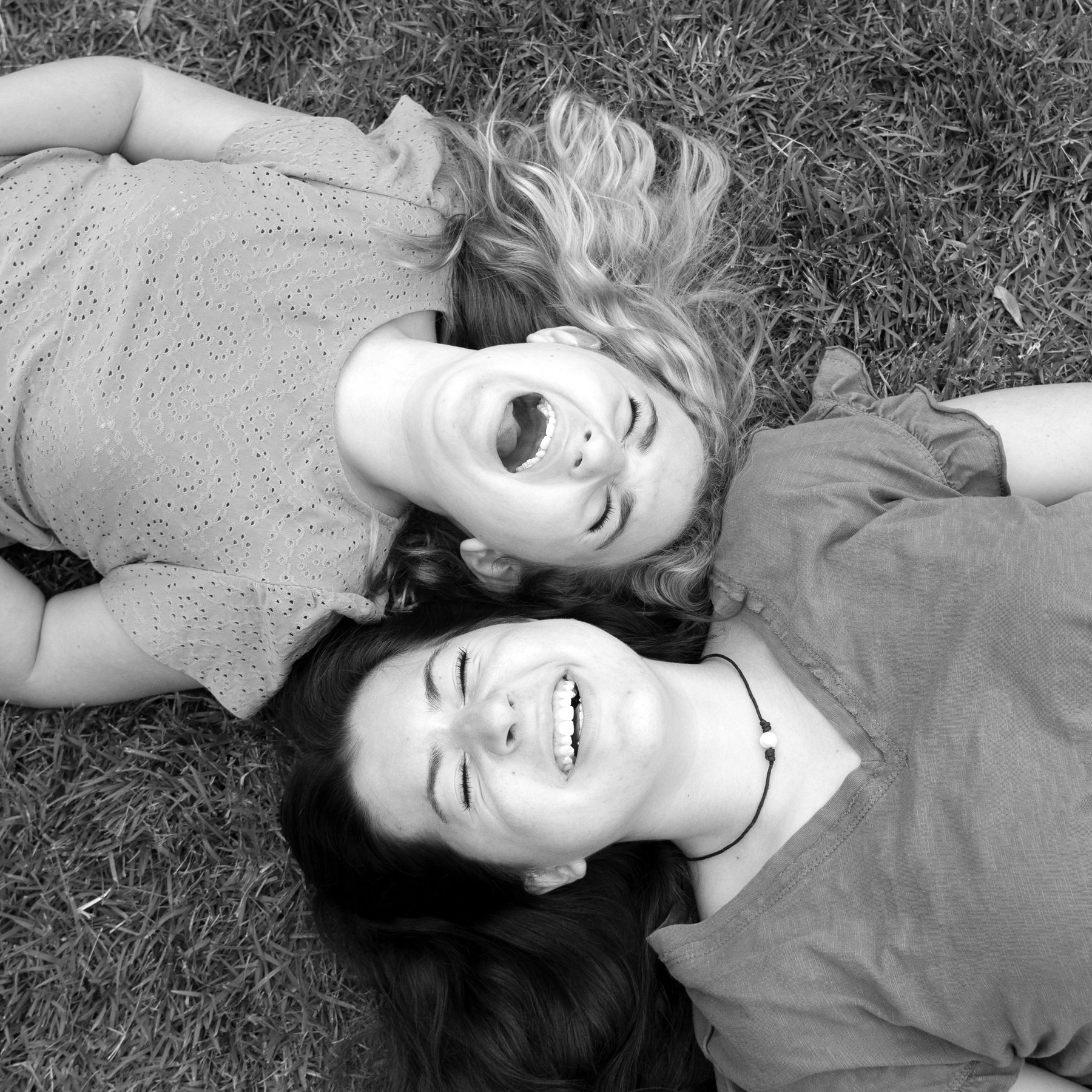 Two young women lying on grass, laughing, with heads close together.
