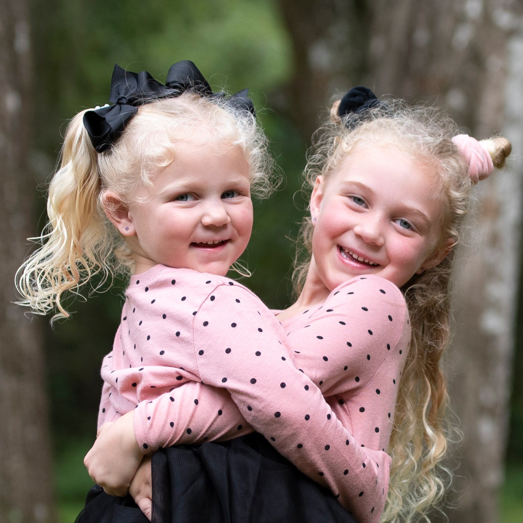 Two young girls with light curly hair, wearing pink polka dot tops, smiling and hugging each other outdoors with trees in the background.