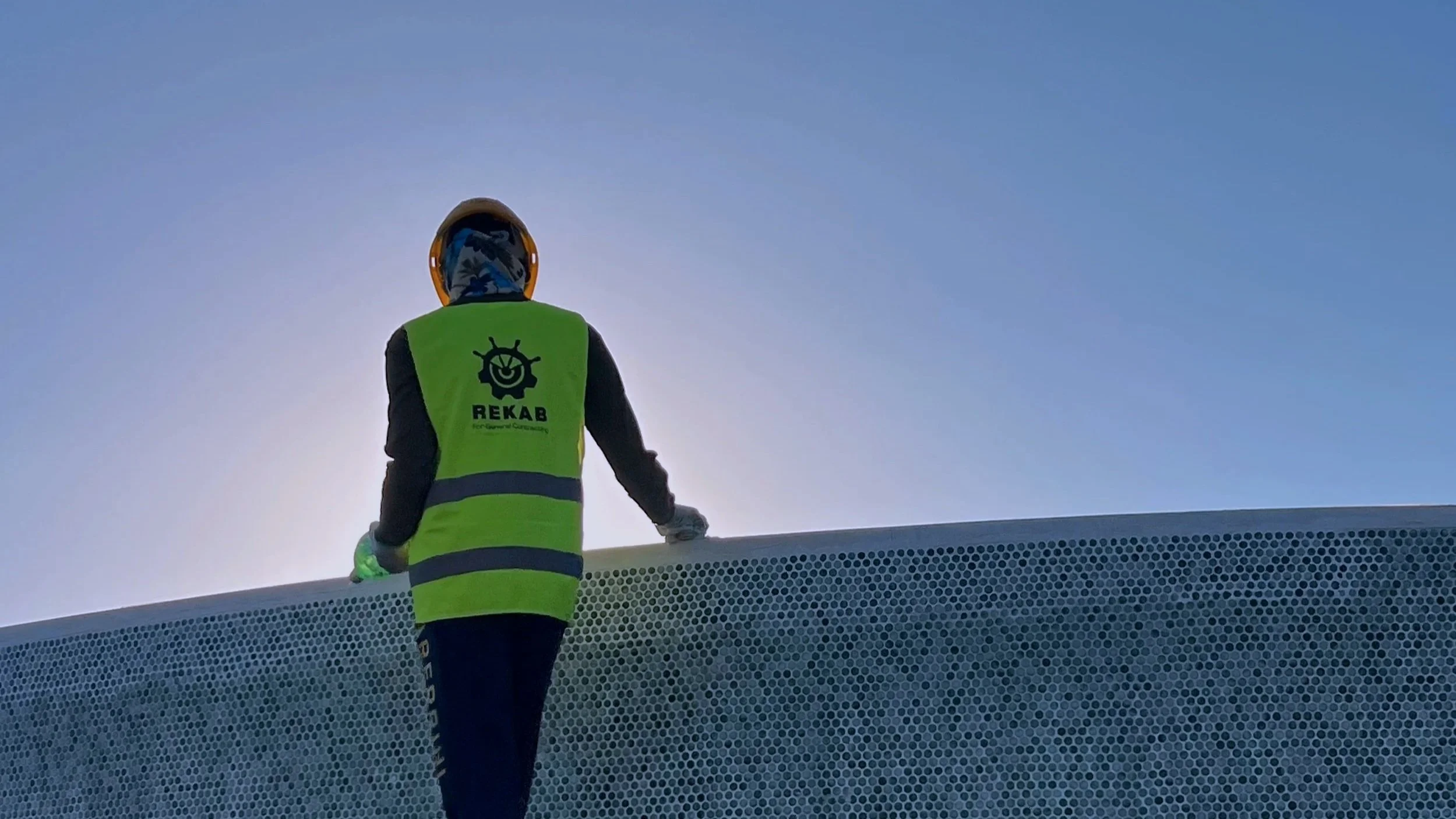 A person wearing a yellow safety helmet and a green safety vest with a logo and the word 'REKAB' stands on top of a large, perforated white surface outdoors, against a clear blue sky.