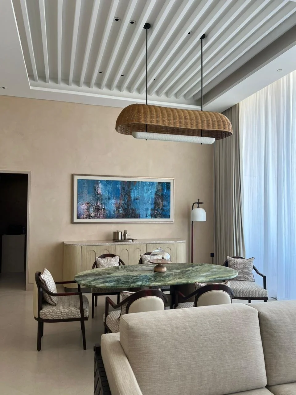 Interior of a modern dining room with a green marble table, six chairs, a beige sofa, a decorative painting, a wooden hanging light fixture, and large sheer curtains.