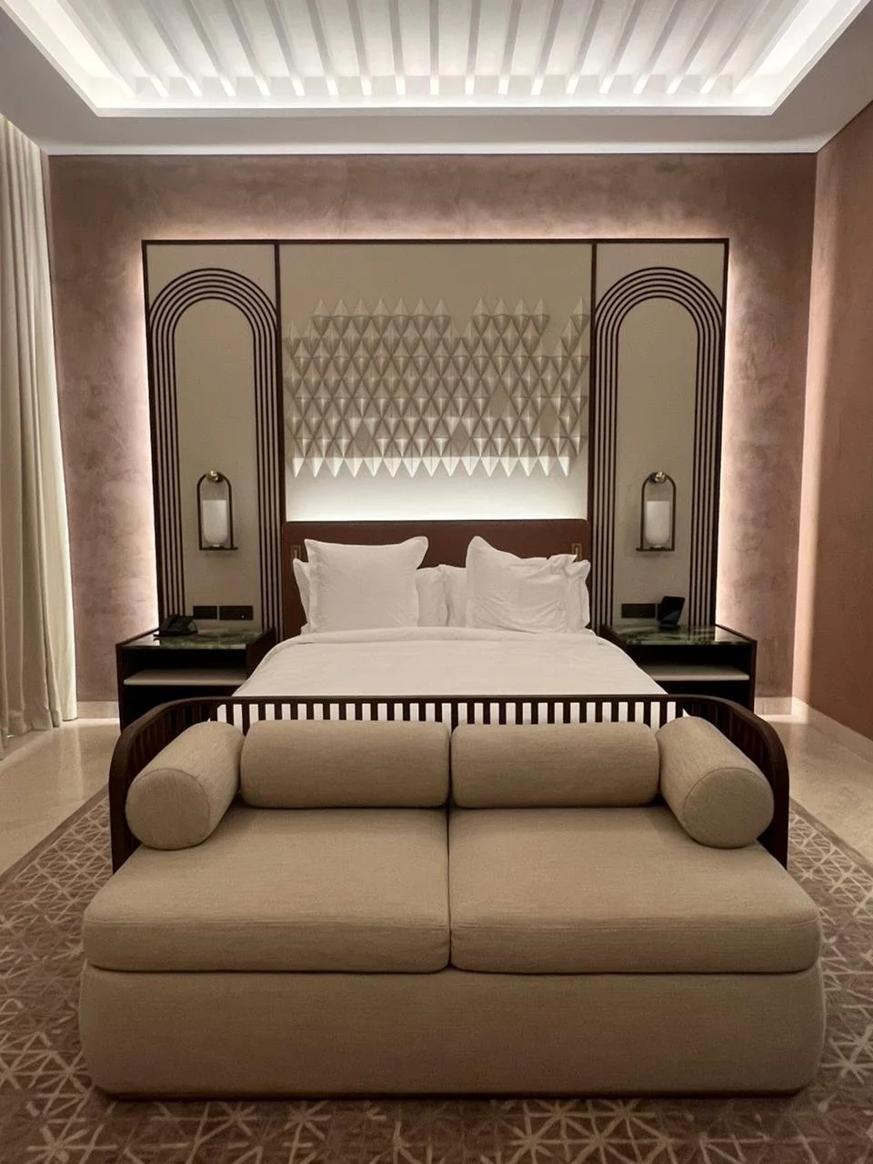 Luxurious hotel room featuring a bed with white bedding, decorative headboard, and bedside lamps. A beige sofa with cylindrical pillows faces the bed, and a patterned carpet completes the elegant decor.