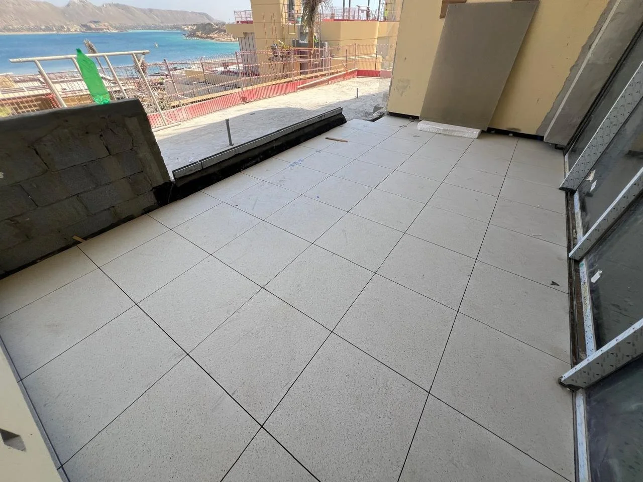 Construction site balcony with white tiled floor overlooking a coastal town and ocean, with construction materials and safety railings.