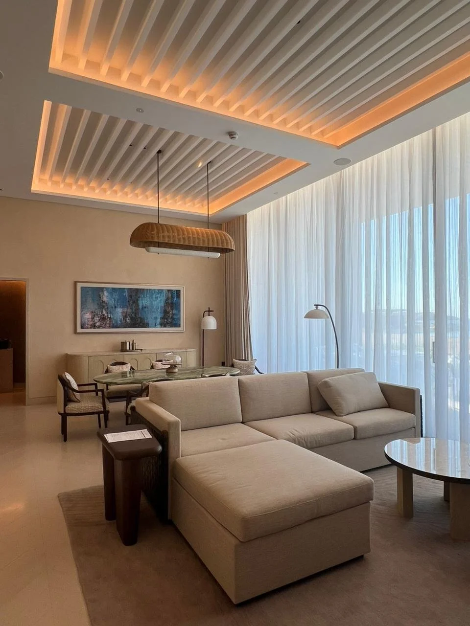 Modern living room with beige sectional sofa, floor lamp, wooden coffee table, and large windows with sheer curtains letting in natural light.
