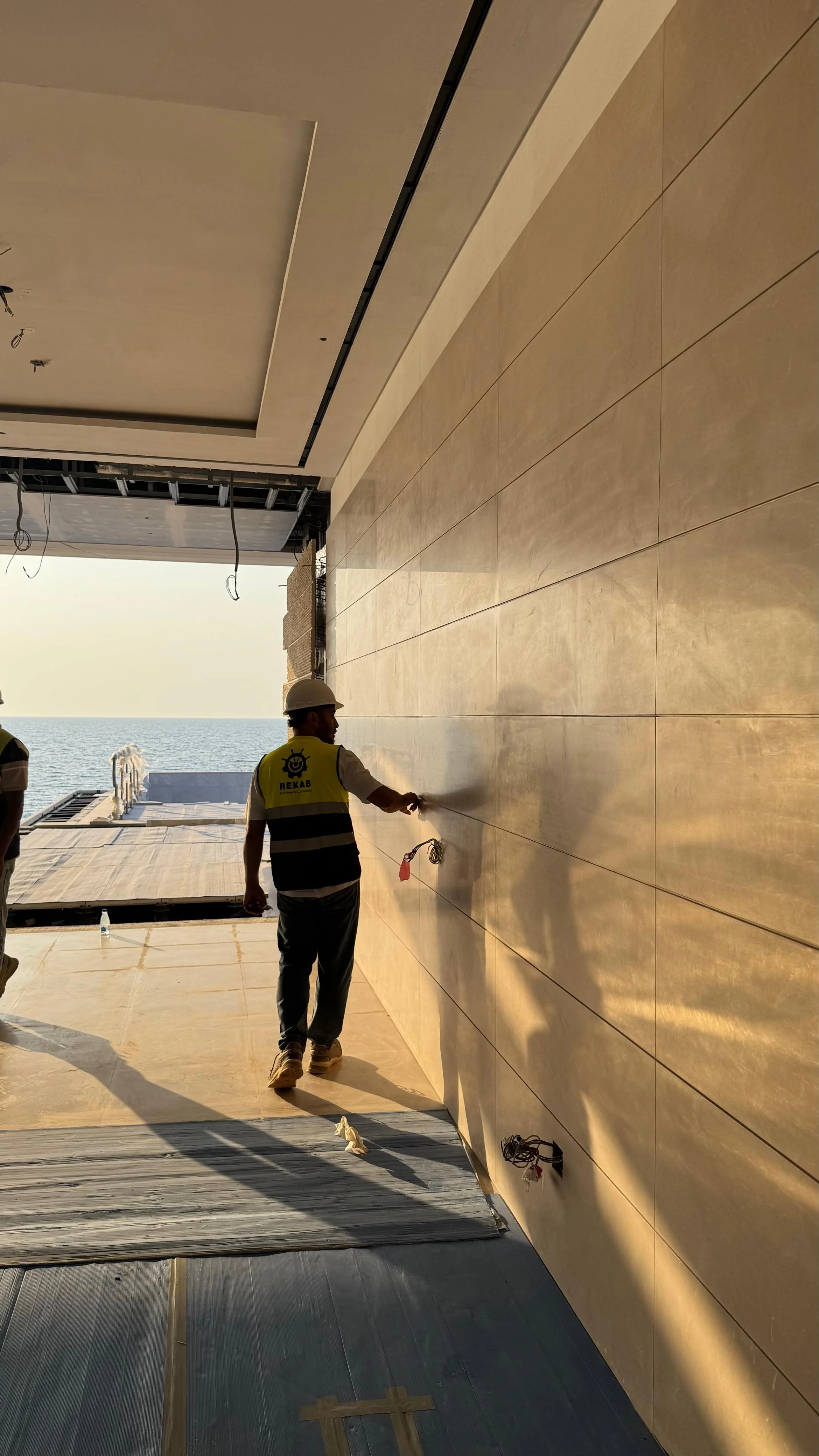 Construction workers installing or cleaning beige wall tiles near the ocean, with one worker spraying the wall as the sun sets.