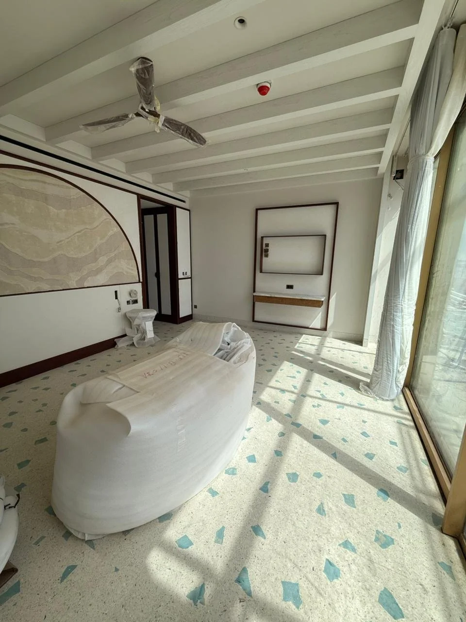 Unoccupied hotel room with a white bed, covered in protective plastic, next to a large window with sheer curtains. The room has terrazzo flooring, and a ceiling fan wrapped in plastic. There is a mounted wall feature and a closet in the corner.