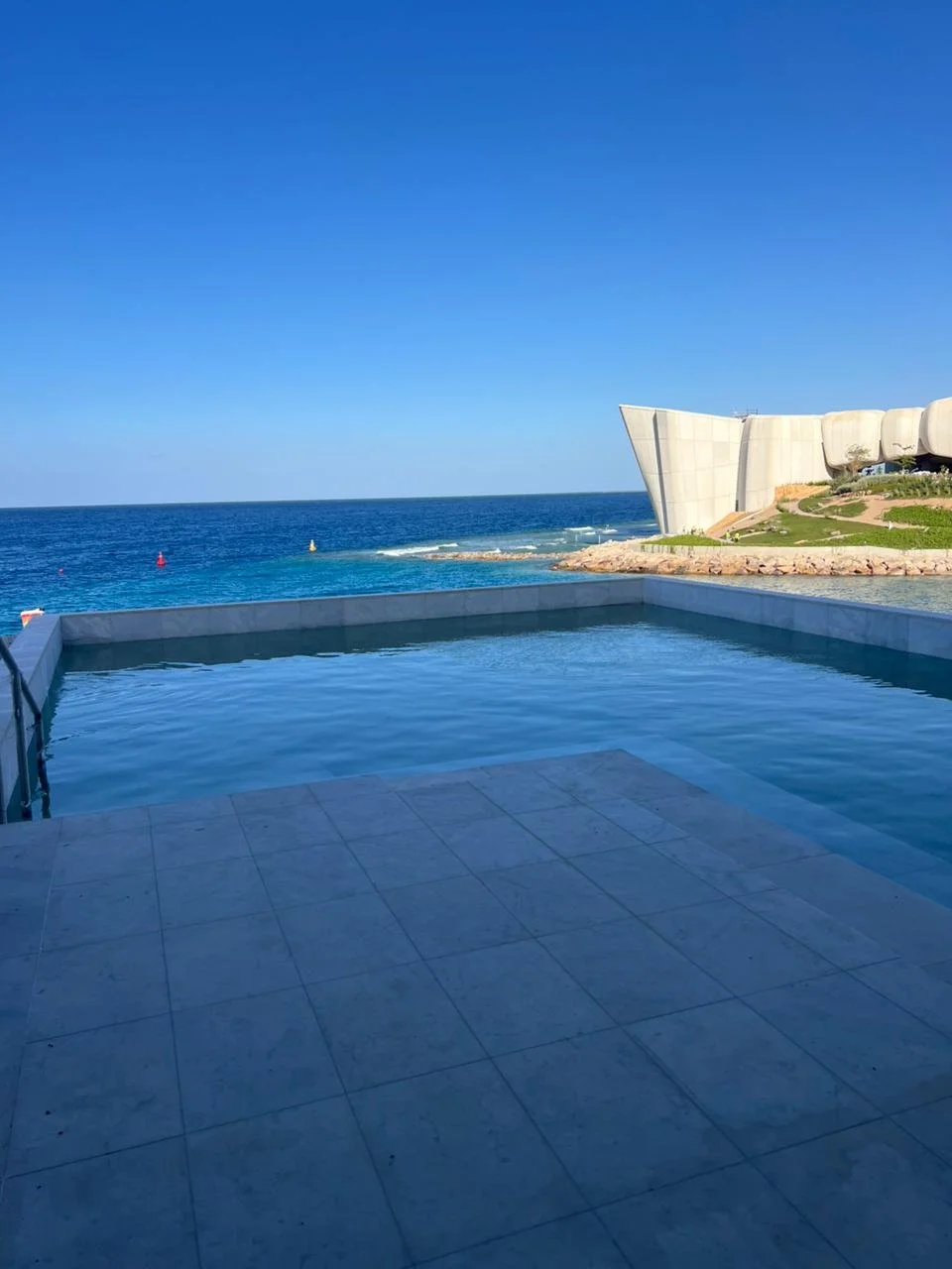 View of a pool with the ocean and a modern building in the background under a clear blue sky.