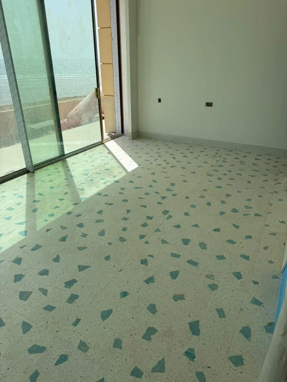 Empty room with terrazzo floor, sunlight coming through glass sliding door, and partially finished wall with exposed electrical outlets.