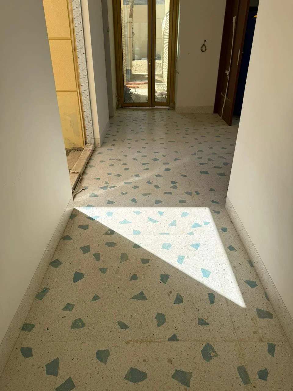 Interior hallway with terrazzo flooring, a sliding glass door at the end leading outside, and a partially open wooden door to the right, with sunlight casting a shadow on the floor.
