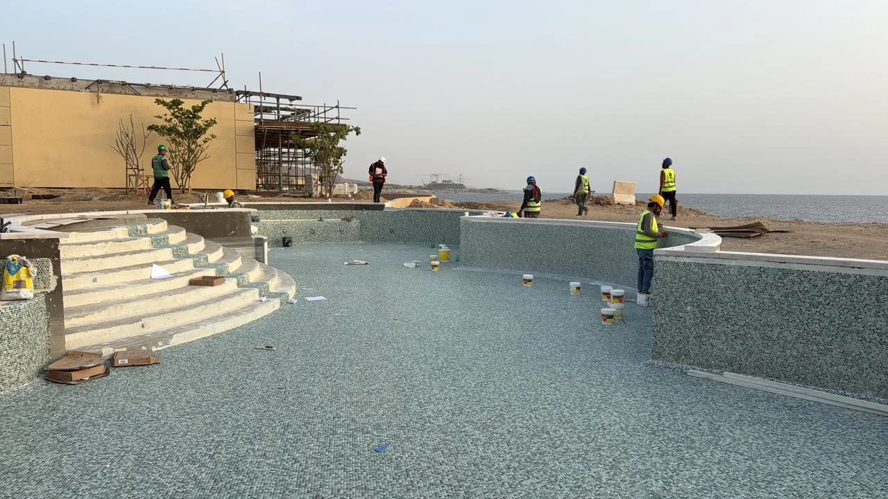Construction workers installing mosaic tiles on a pool near the beach, with scaffolding and a building under construction in the background.