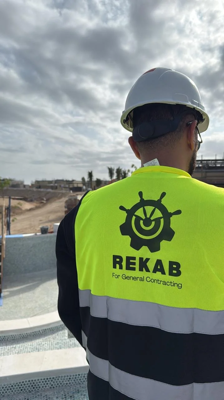 A construction worker wearing a white helmet and a high-visibility yellow vest with black and white stripes, standing outdoors at a construction site with a cloudy sky in the background.