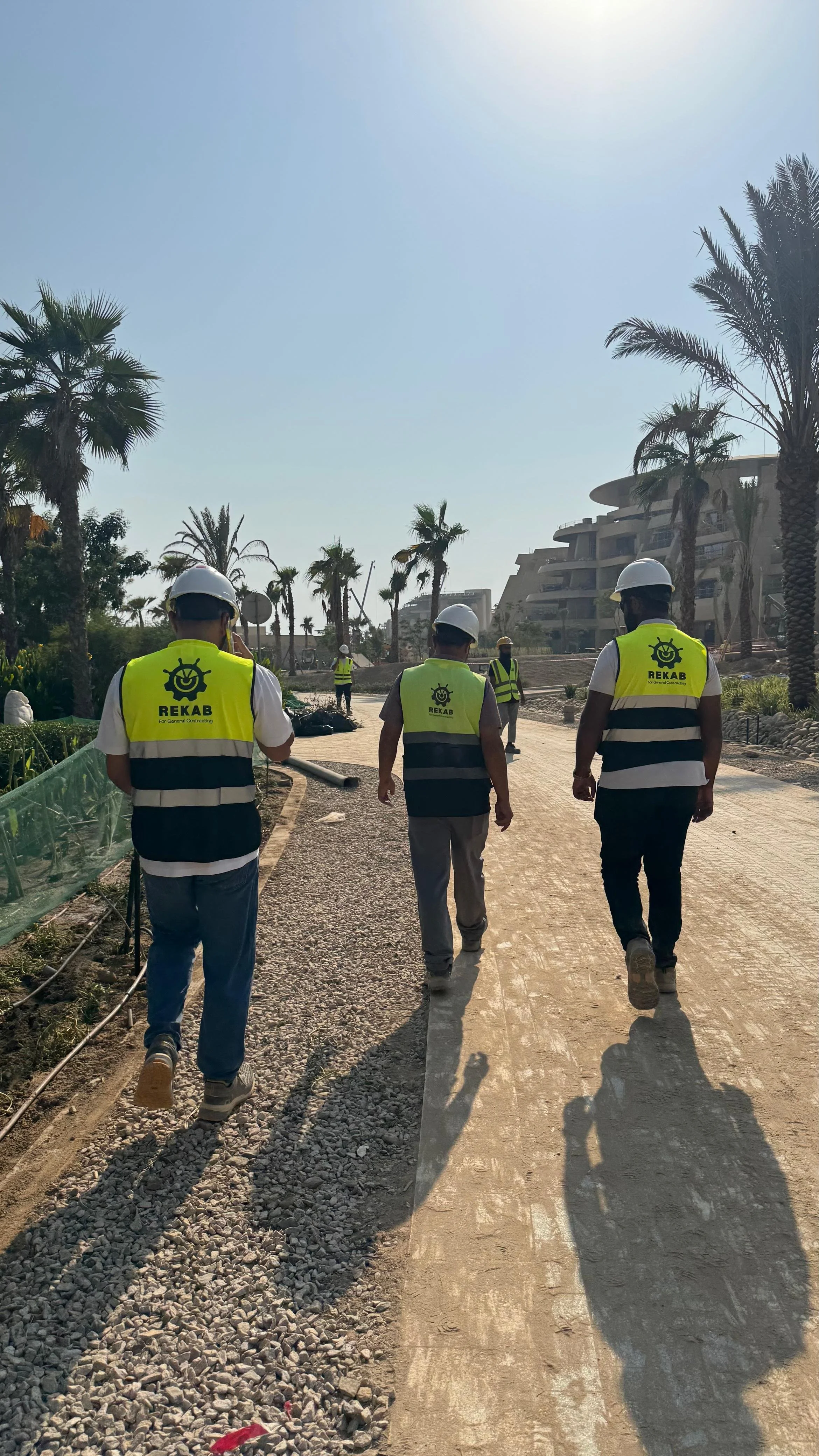 Construction workers walking on a partially paved pathway on a sunny day, surrounded by palm trees and modern buildings in the background.