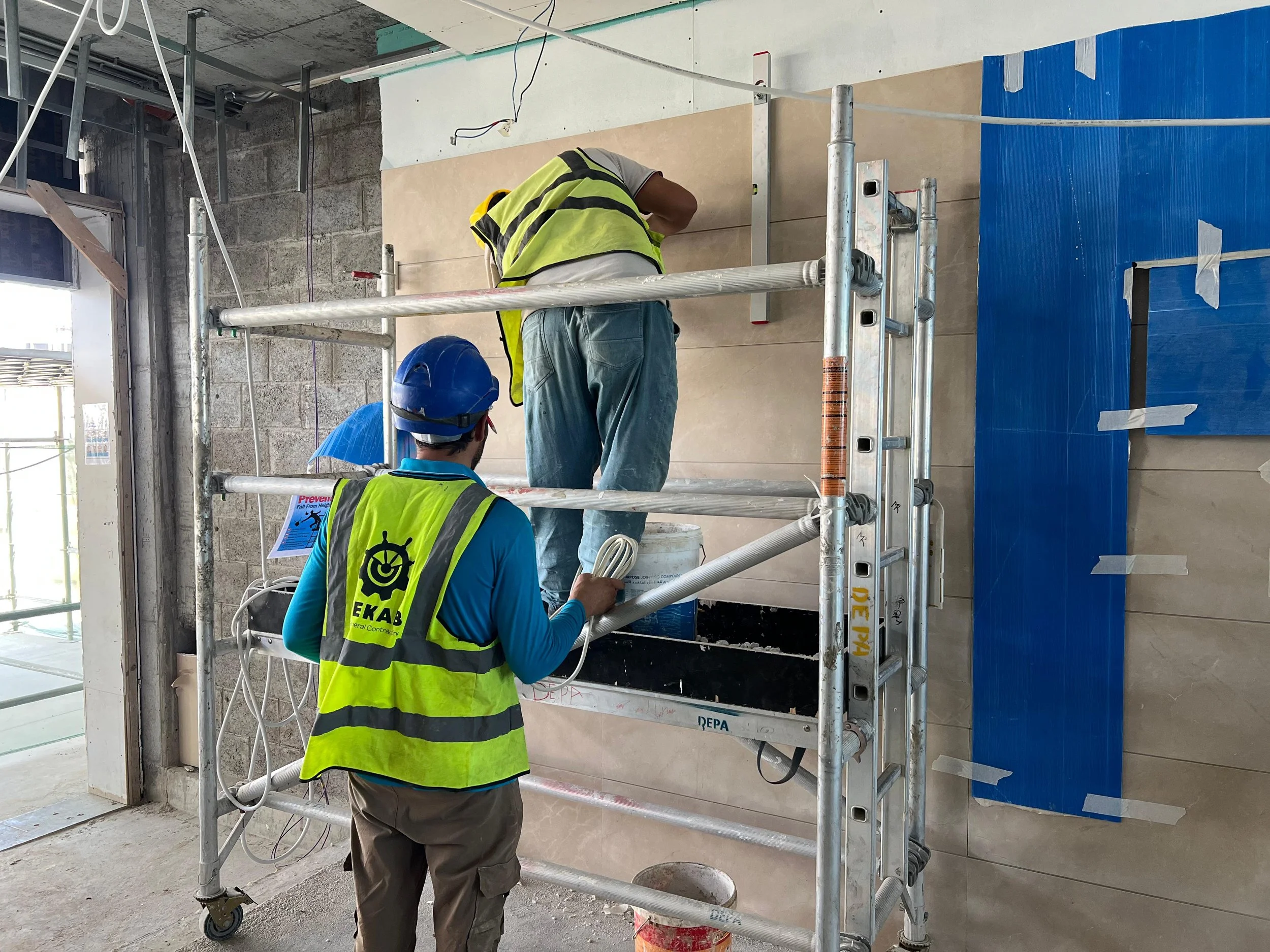 Two construction workers wearing safety vests and helmets working on a scaffold on a wall in an unfinished building.