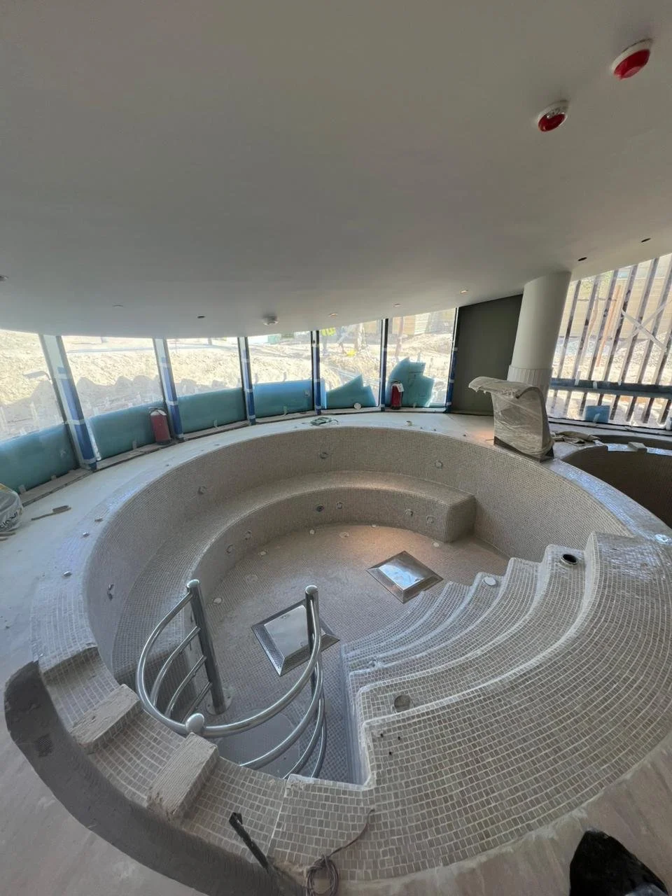 Under construction indoor hot tub or small pool area with tiled steps and rounded seating area, surrounded by large windows, construction materials, and safety barriers.