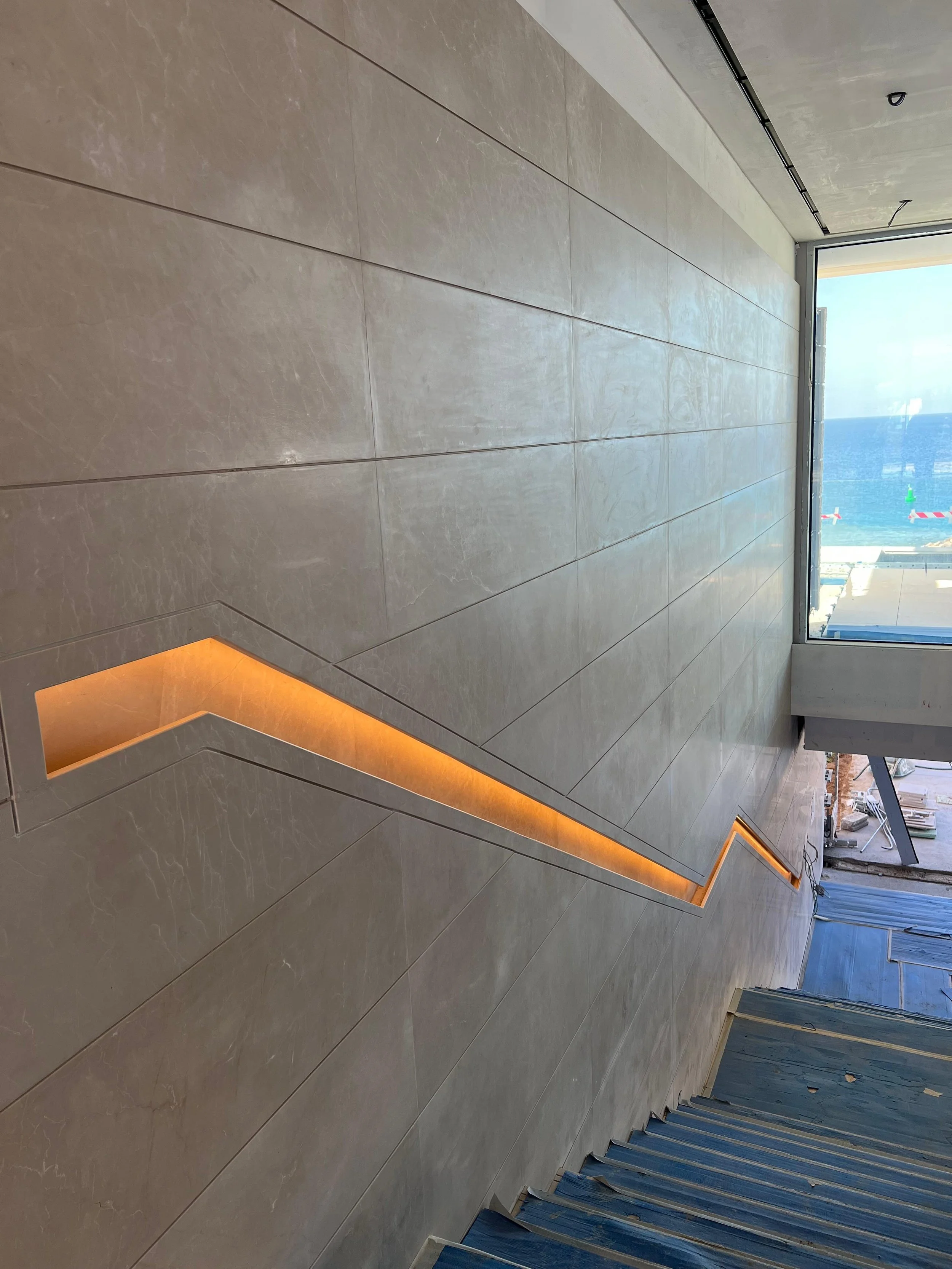 Interior view of a modern staircase with beige tiled walls and a zigzag-shaped light strip built into the wall, overlooking a large window with a view of the ocean and outdoor construction.