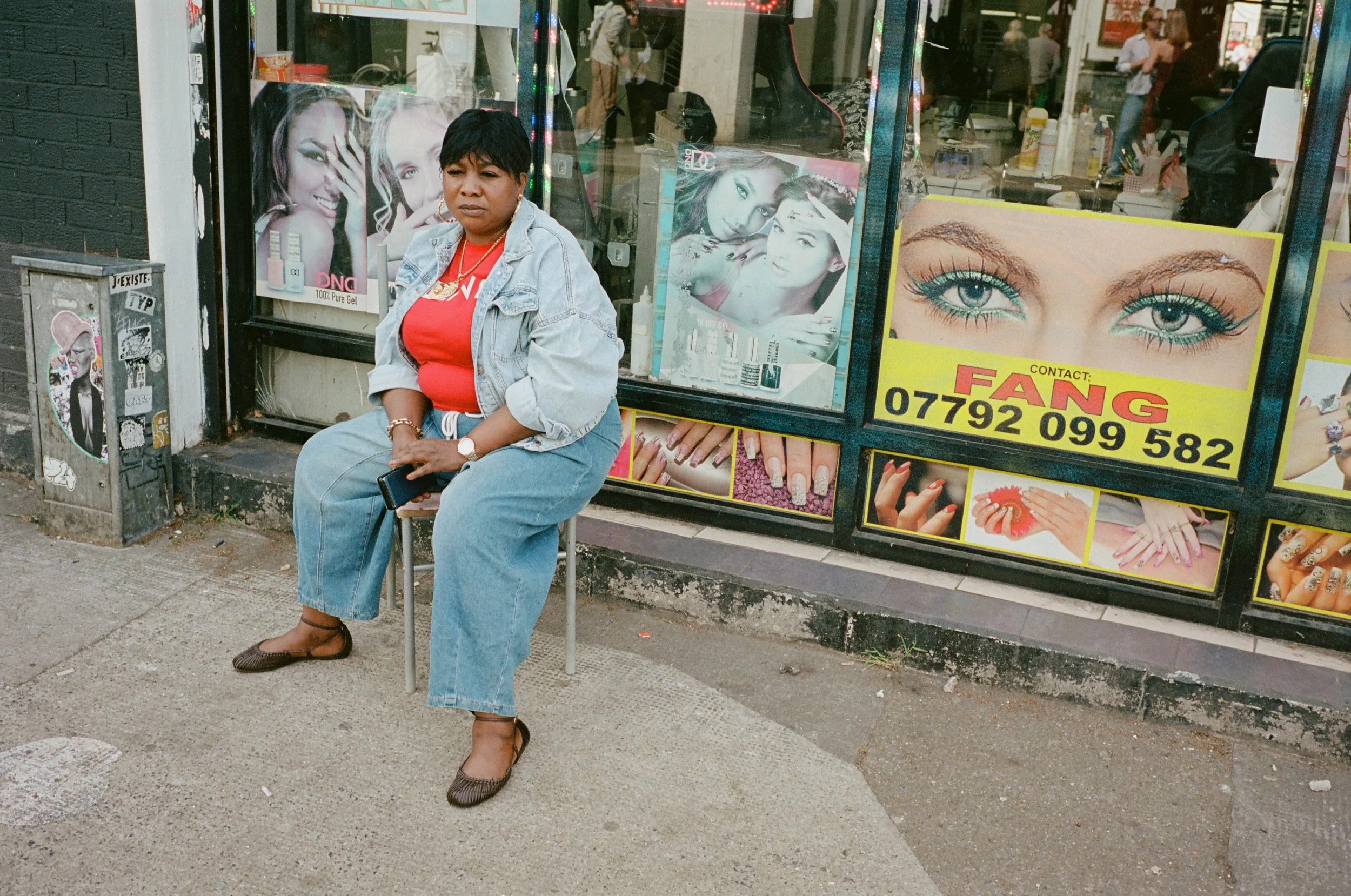 A woman sitting on a small metal chair outside a beauty shop with colorful signs and posters of makeup, nails, and beauty products in the window.