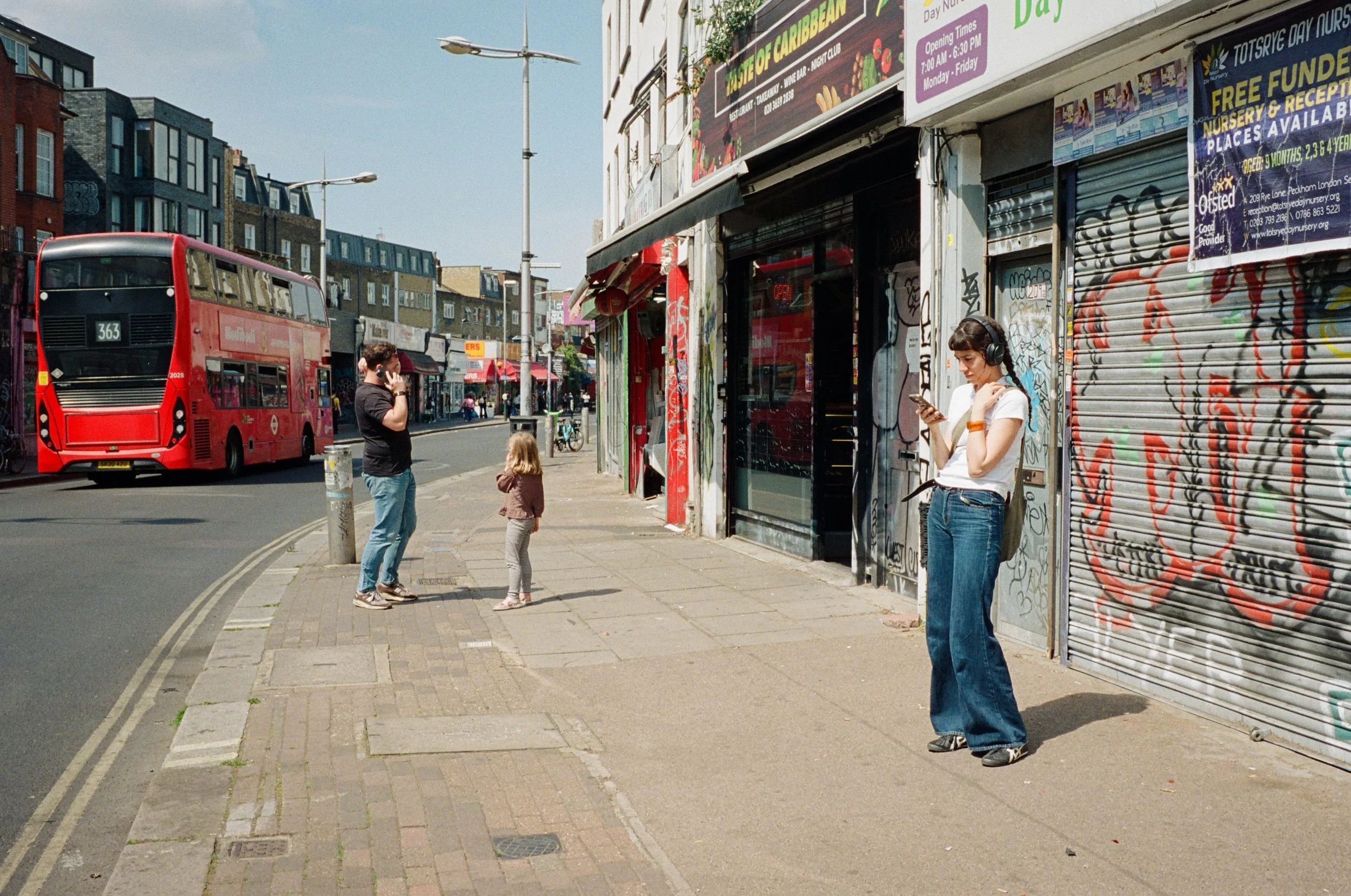 People standing on a city sidewalk near a graffiti-covered shopfront, with a red double-decker bus passing by and residential buildings in the background.
