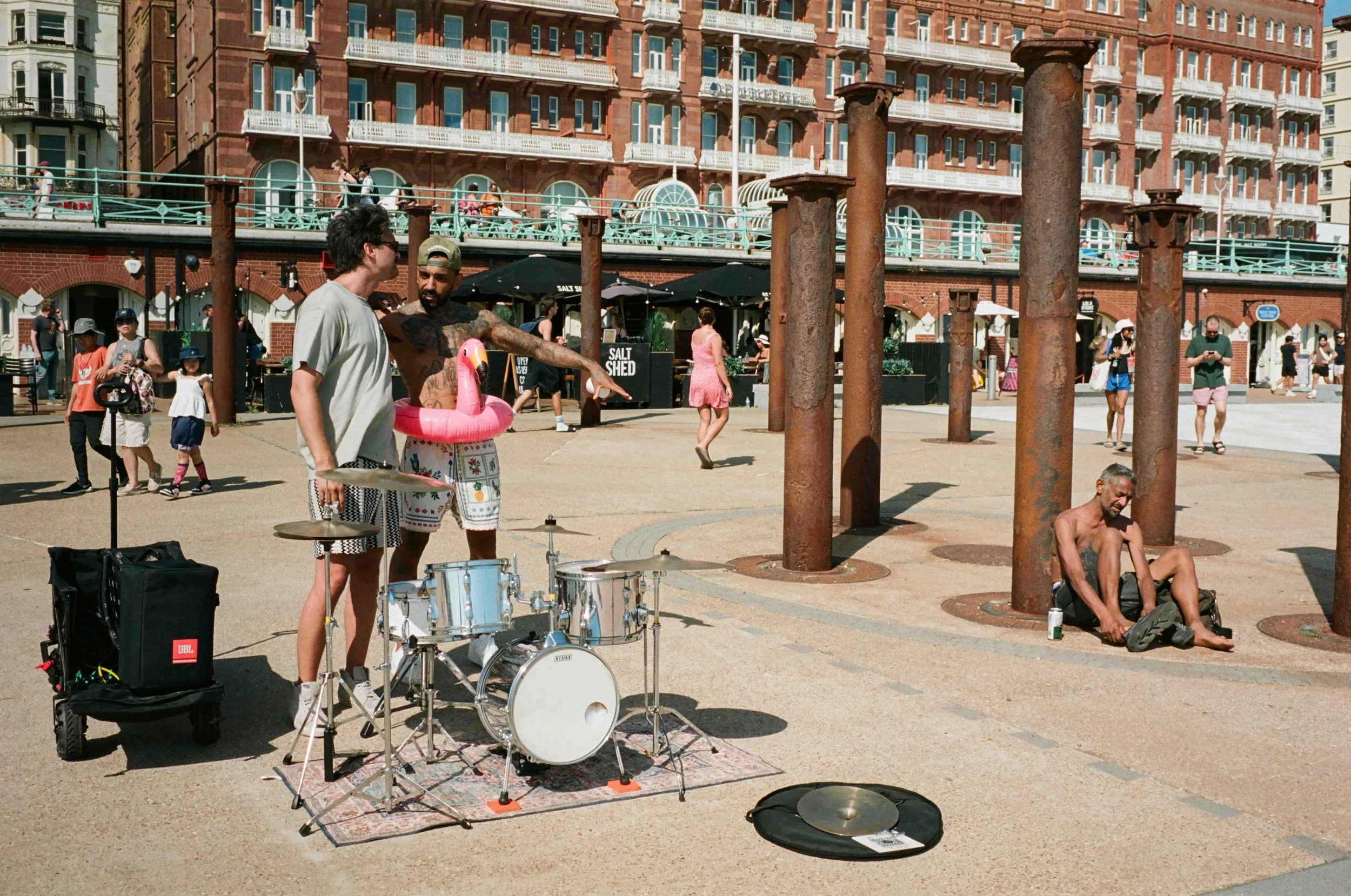 Street scene with musicians playing drums and a man sitting on the ground, rusted columns, people walking, and a historic brick building with balconies and blue railings in the background.
