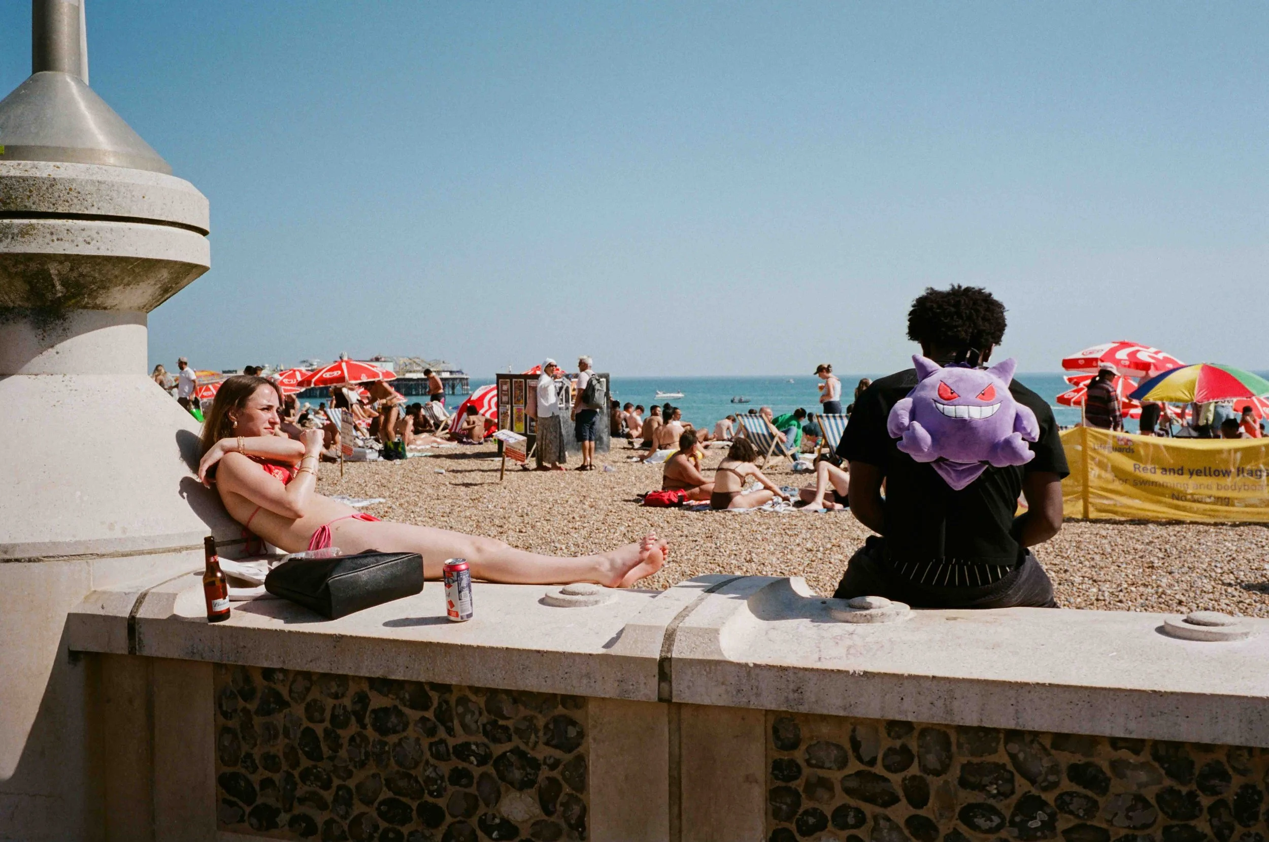 A crowded beach scene with people relaxing, sunbathing, and walking near the ocean under colorful umbrellas. In the foreground, a young woman with long hair is sitting on concrete, leaning against a wall, with a beer and soda can nearby. A person wit