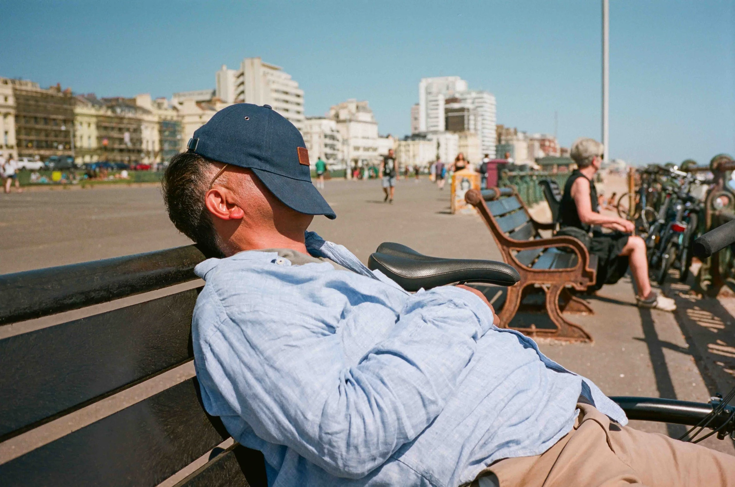 A man is sleeping on a park bench near an urban area with tall buildings in the background. He is wearing a blue cap and a light-colored hoodie.