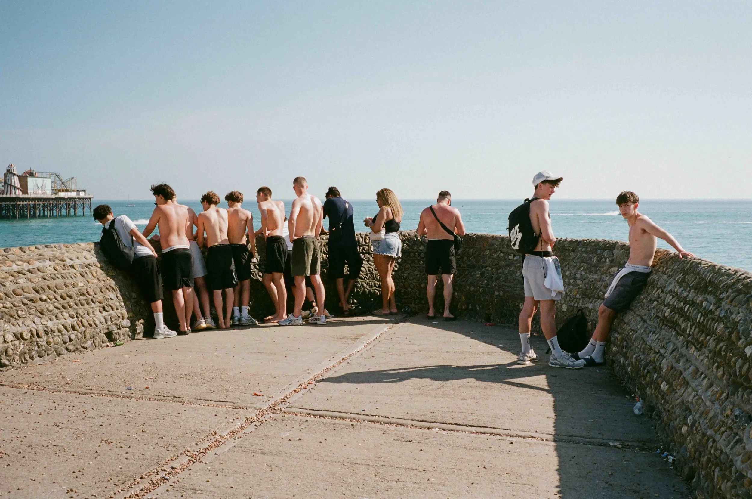Group of young men and women standing and sitting by a stone wall near the ocean, some shirtless, some with backpacks, in a sunny setting.