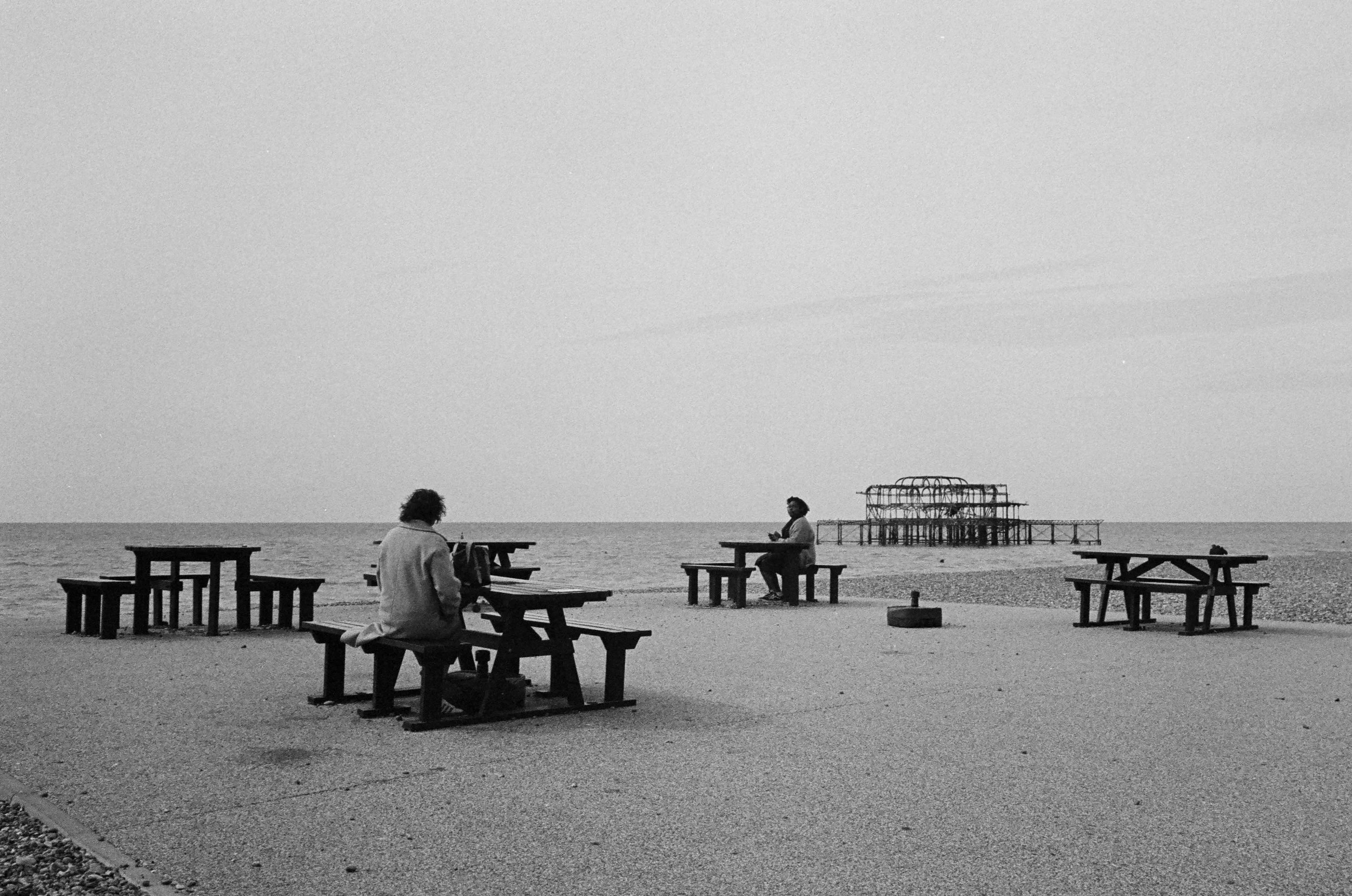 Black and white photo of a beach with two people sitting at picnic tables, looking at the ocean. In the background, there is a partially submerged, old pier structure.