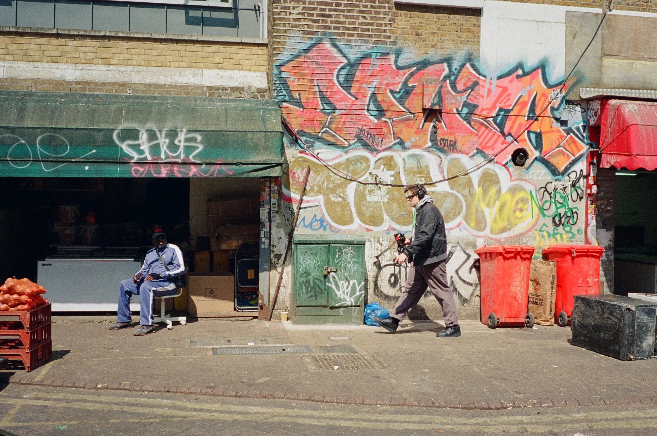 Street scene featuring two men in front of a building with colorful graffiti. One man is sitting on a bench wearing blue work clothes and a red cap, the other man is walking past wearing dark clothes and headphones. There are red and green trash bins