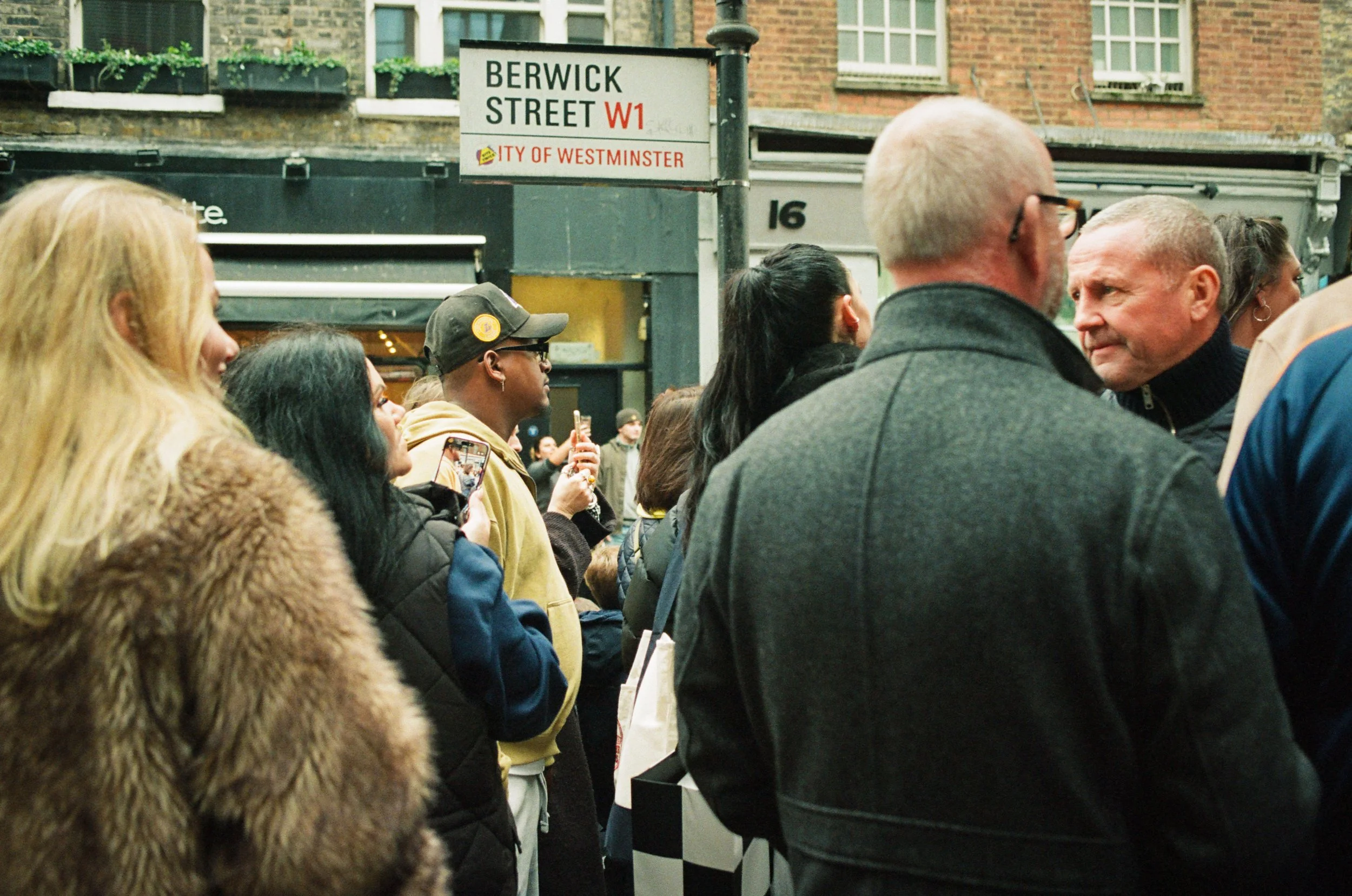 Crowd of people standing in line on Berwick Street in Westminster, London, England, with some individuals looking at their phones and a man holding a drink. Street sign indicating location at the top of the image.