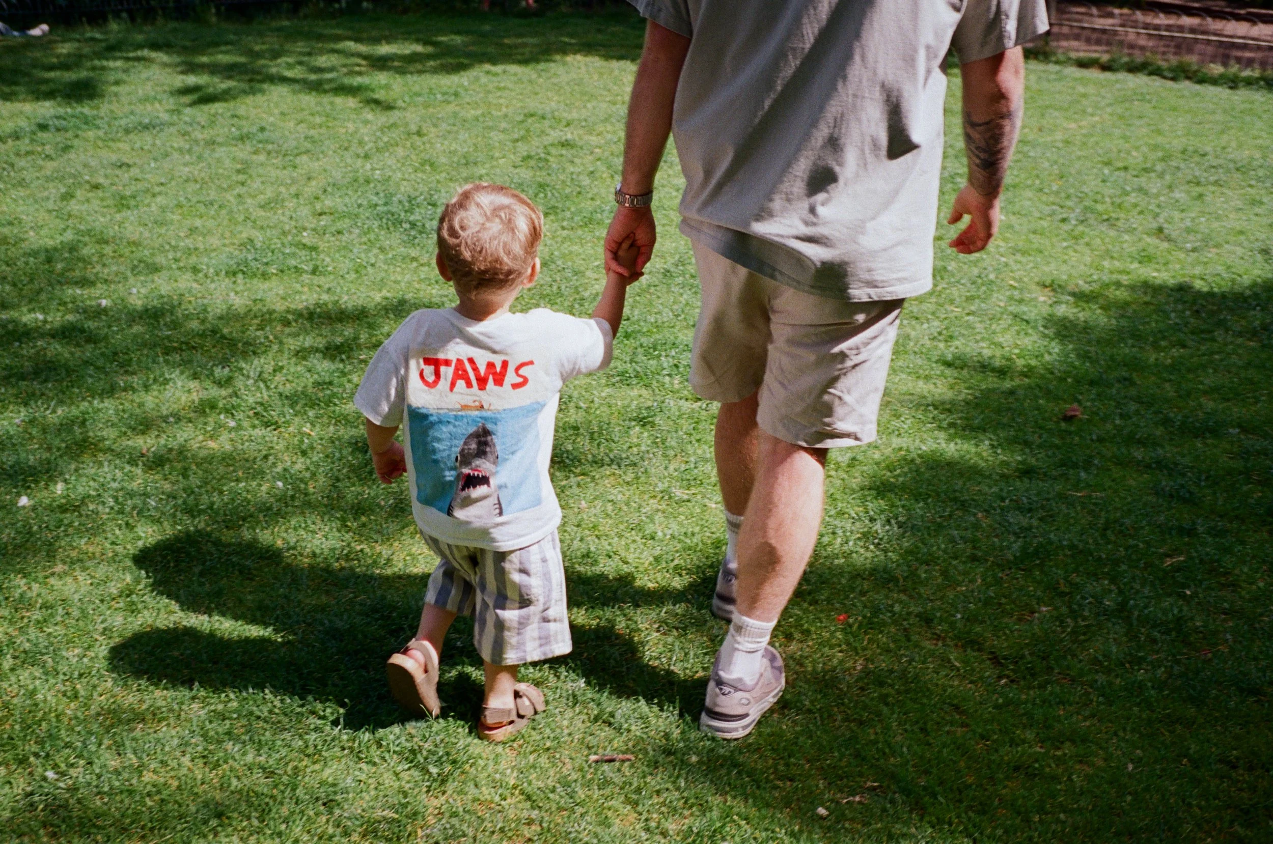 A child holding hands with an adult while walking on a grassy yard, both dressed casually, with the child wearing a Jurassic Park T-shirt and striped shorts.