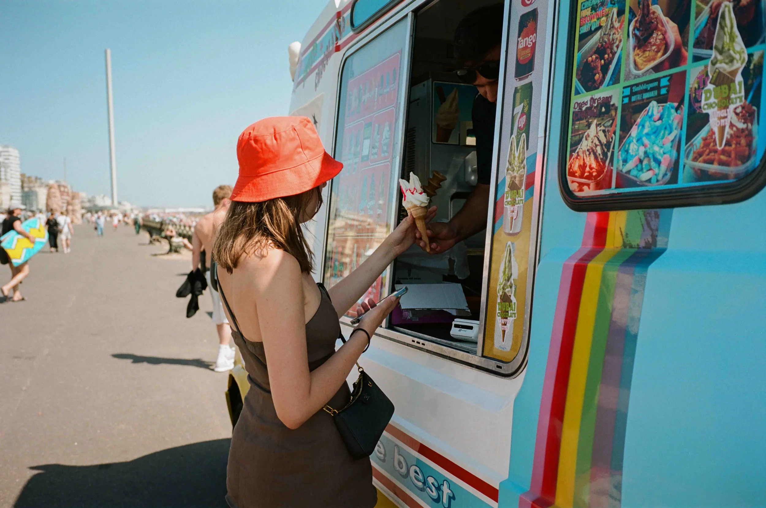 A woman in a black dress and red bucket hat ordering ice cream from a colorful ice cream truck at the beach.