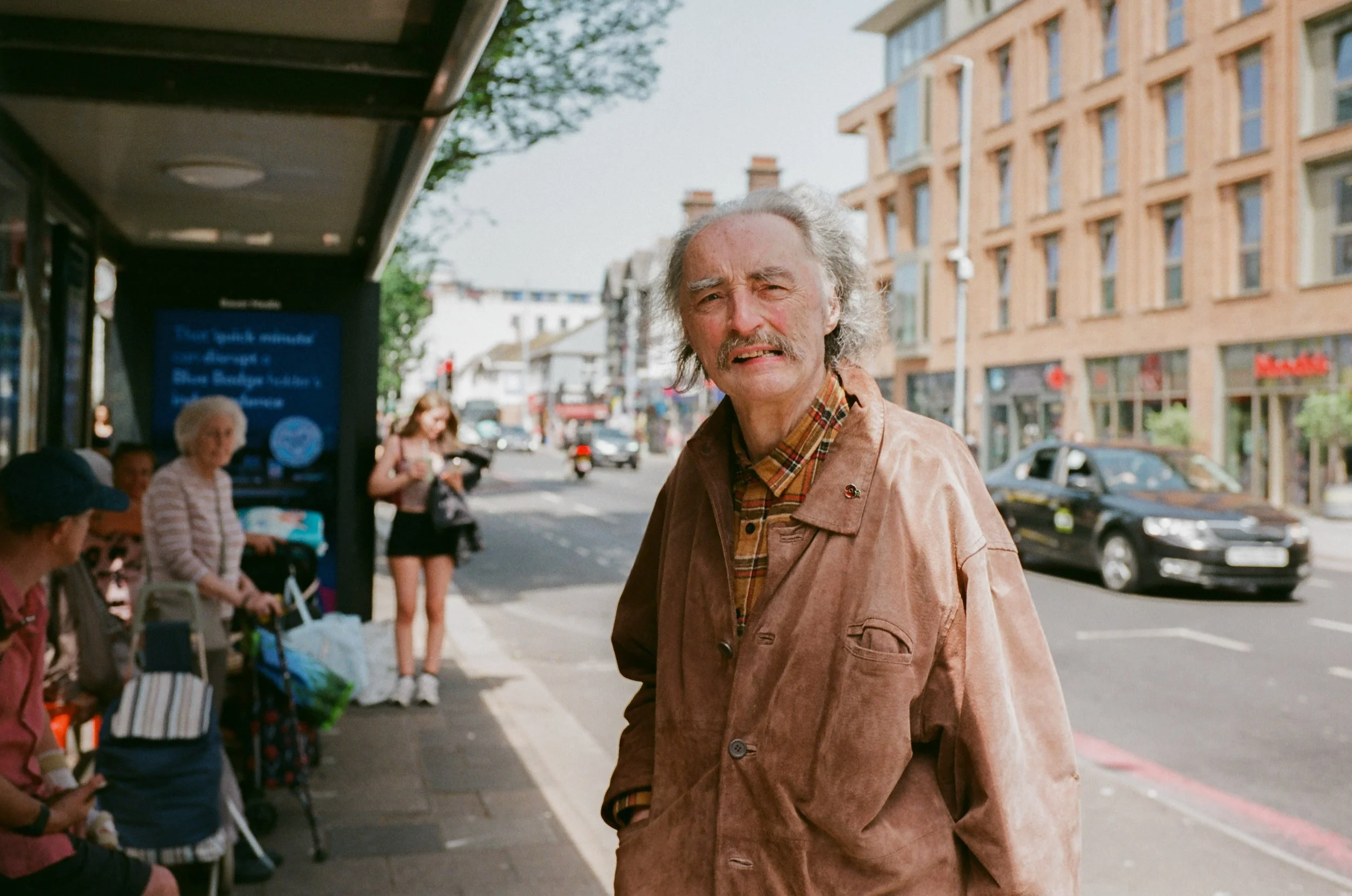 An elderly man with gray hair, a mustache, and wearing a brown jacket over a plaid shirt stands on a city sidewalk, looking directly at the camera. In the background, there are people sitting at a bus stop, a woman standing, and buildings with large 