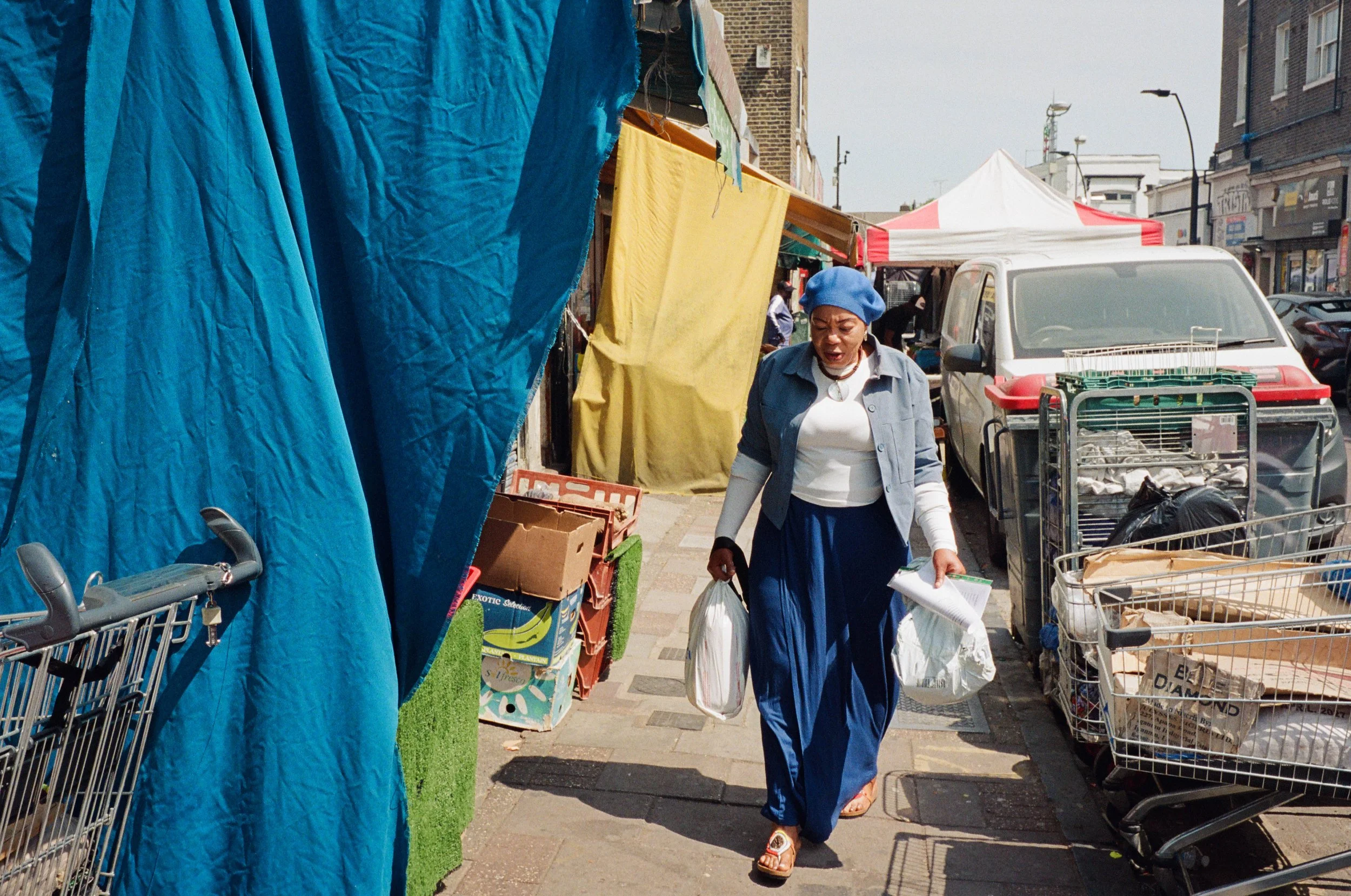 A woman in a blue headscarf and skirt walking through an outdoor market, surrounded by shopping carts and colorful cloths hanging on display.
