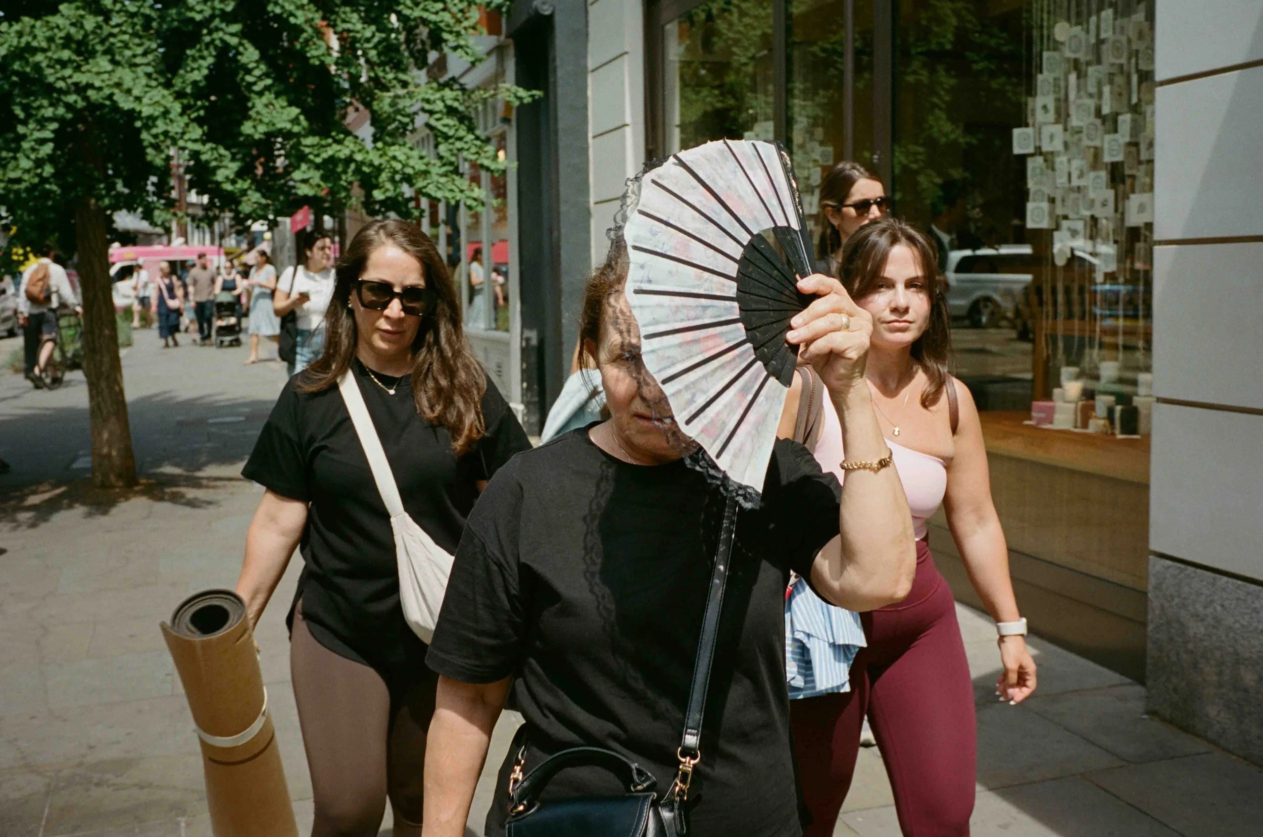 Four women walking on a city sidewalk, one holding a decorative fan in front of her face, with a background of pedestrians, trees, and storefronts.