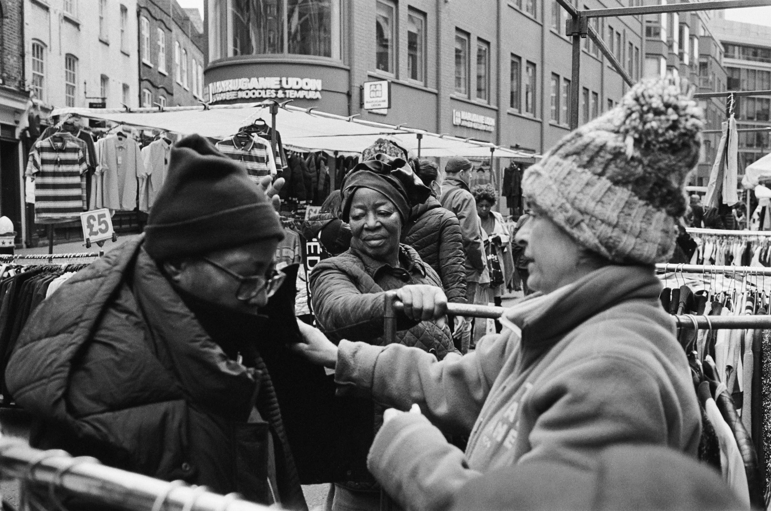 People standing in a busy outdoor market, shopping for clothes and accessories, with stores and signs visible in the background.