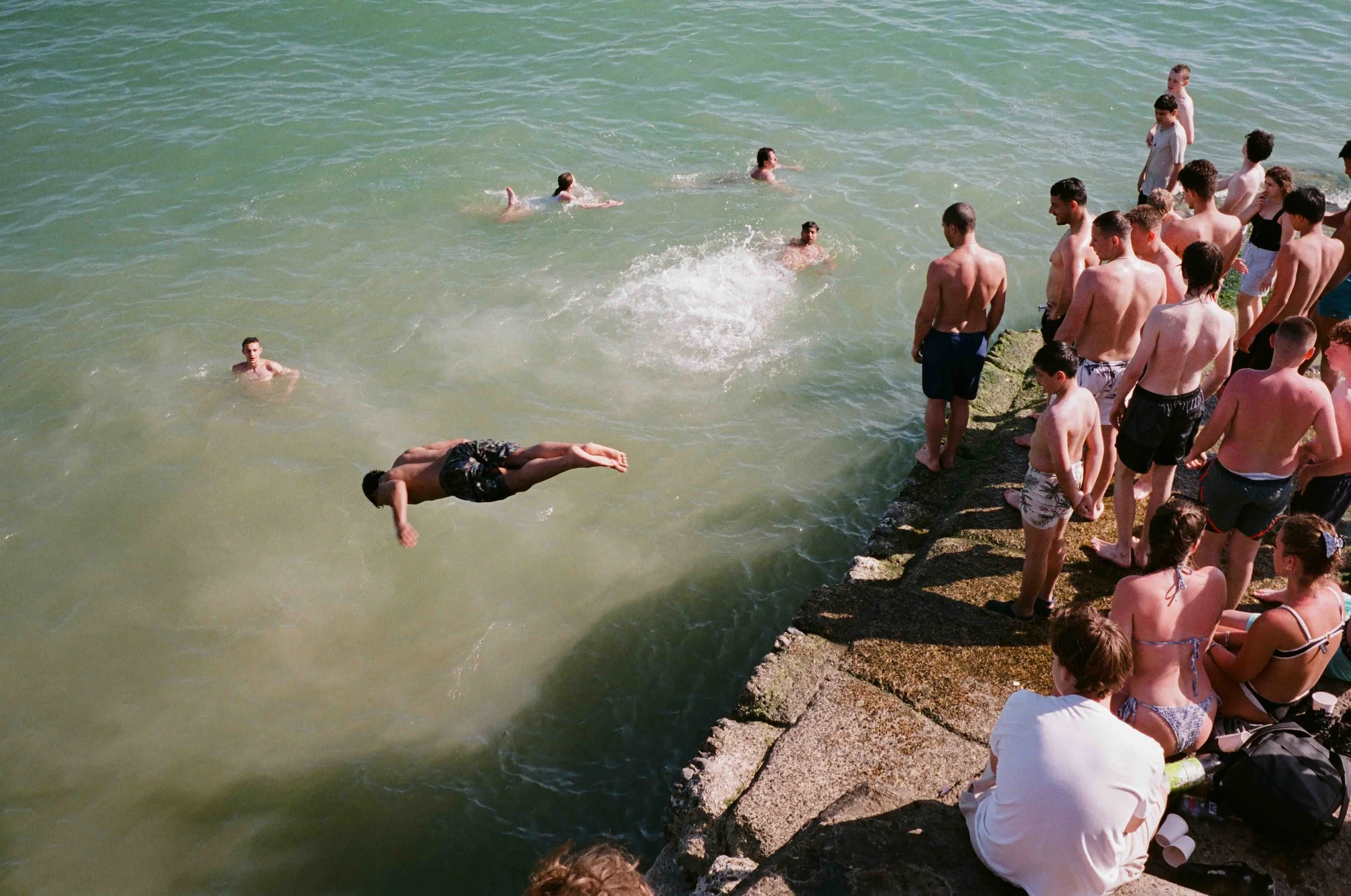 Group of people at the beach, with some swimming in the water and others sitting on rocks along the shoreline.