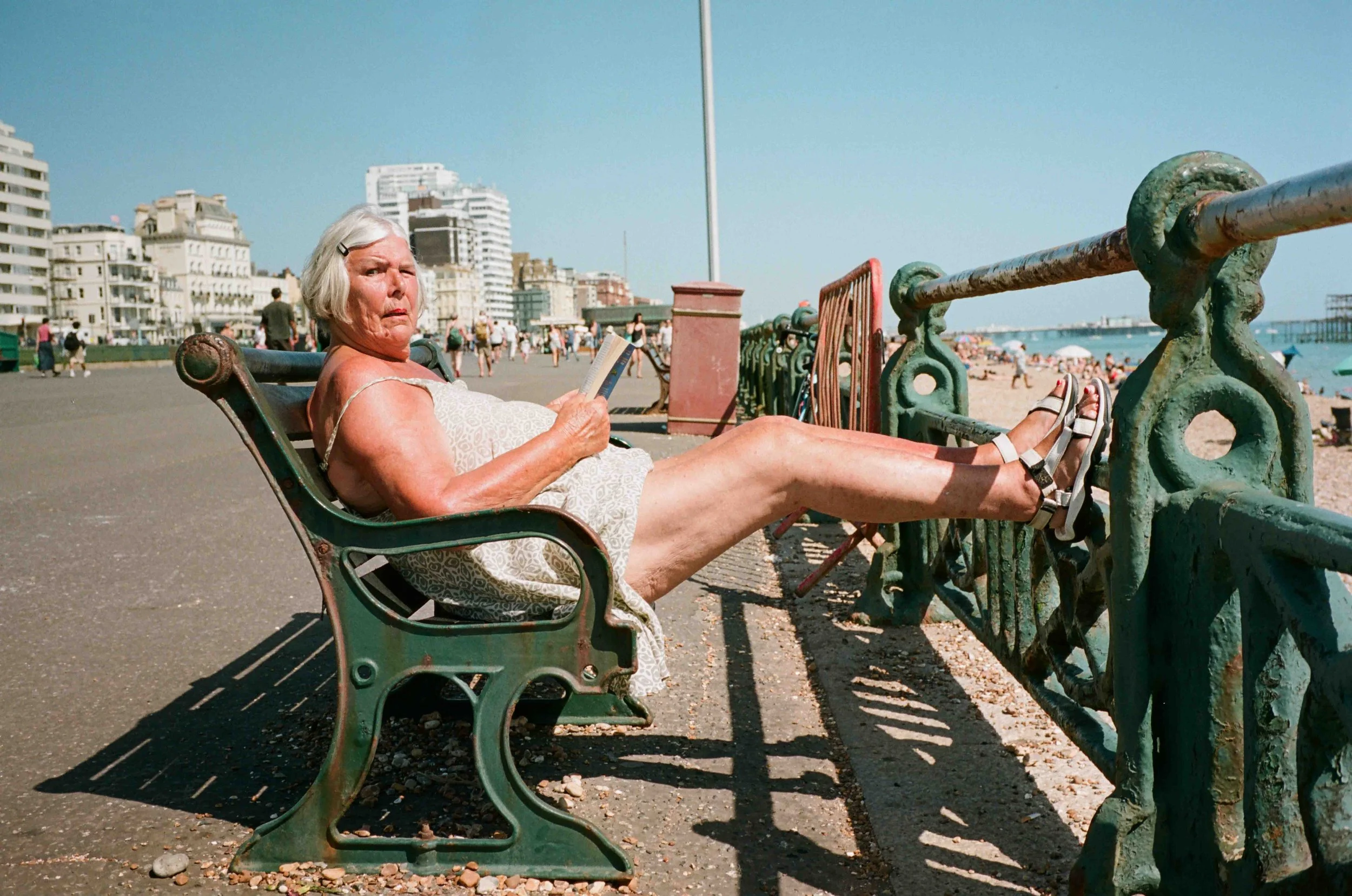 An elderly woman with gray hair, wearing a light-colored dress, is relaxing on a park bench by the beach, reading a book on a sunny day. There are many people walking and enjoying the beach in the background, with tall buildings and clear blue sky vi